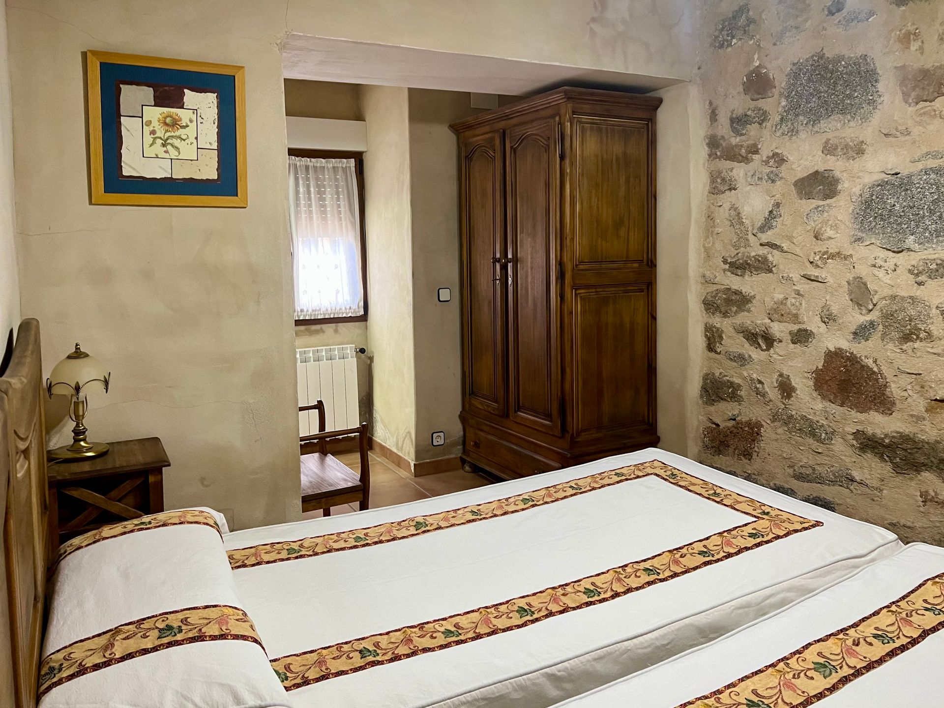 Cozy bedroom with a white bedspread, wooden wardrobe, stone accent wall, and framed art.