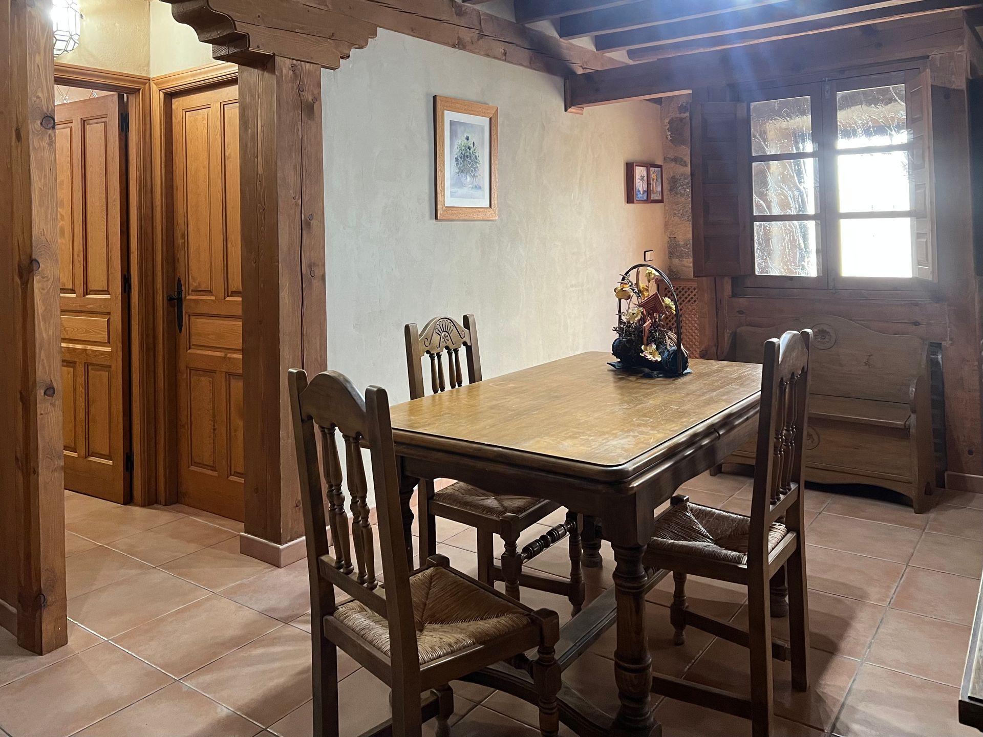Rustic dining room with wooden table, chairs, and beamed ceiling by a window.