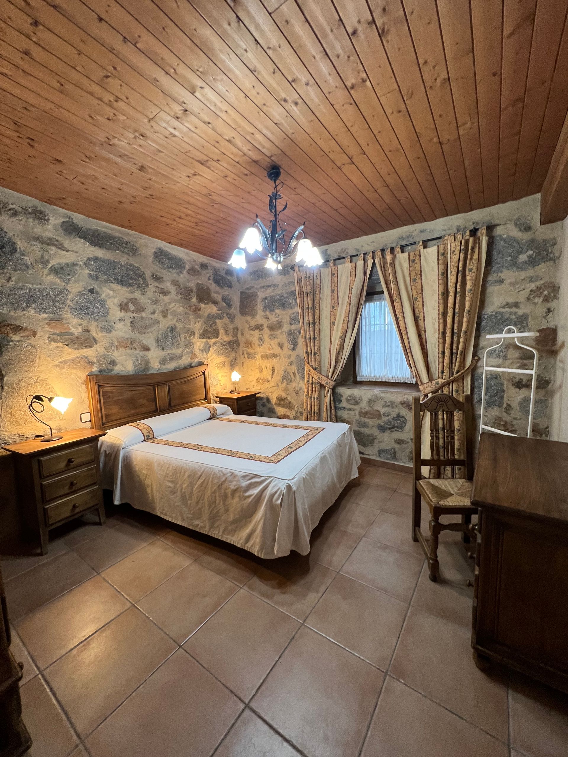 Rustic stone bedroom with wood ceiling, double bed, chandelier, and window with curtains