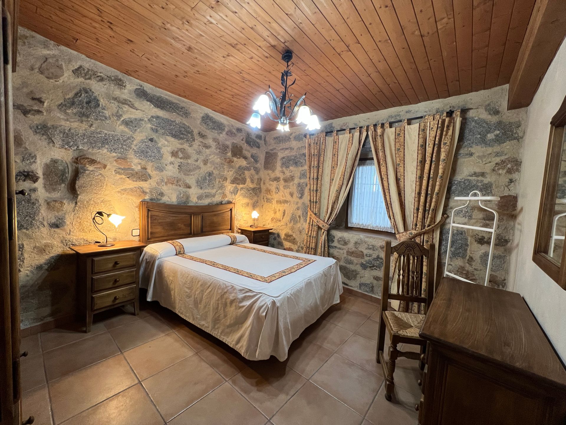Stone-walled bedroom with wooden ceiling, double bed, bedside lamps, desk, and curtained window