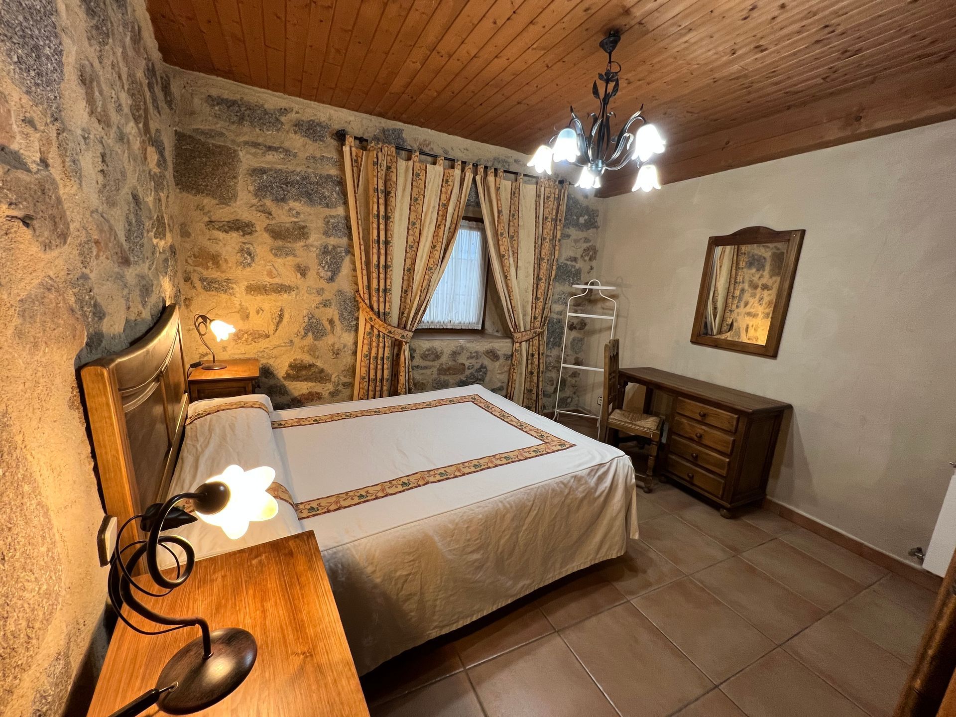 Rustic bedroom with stone walls, wooden ceiling, double bed, dresser, and warm lamp lighting
