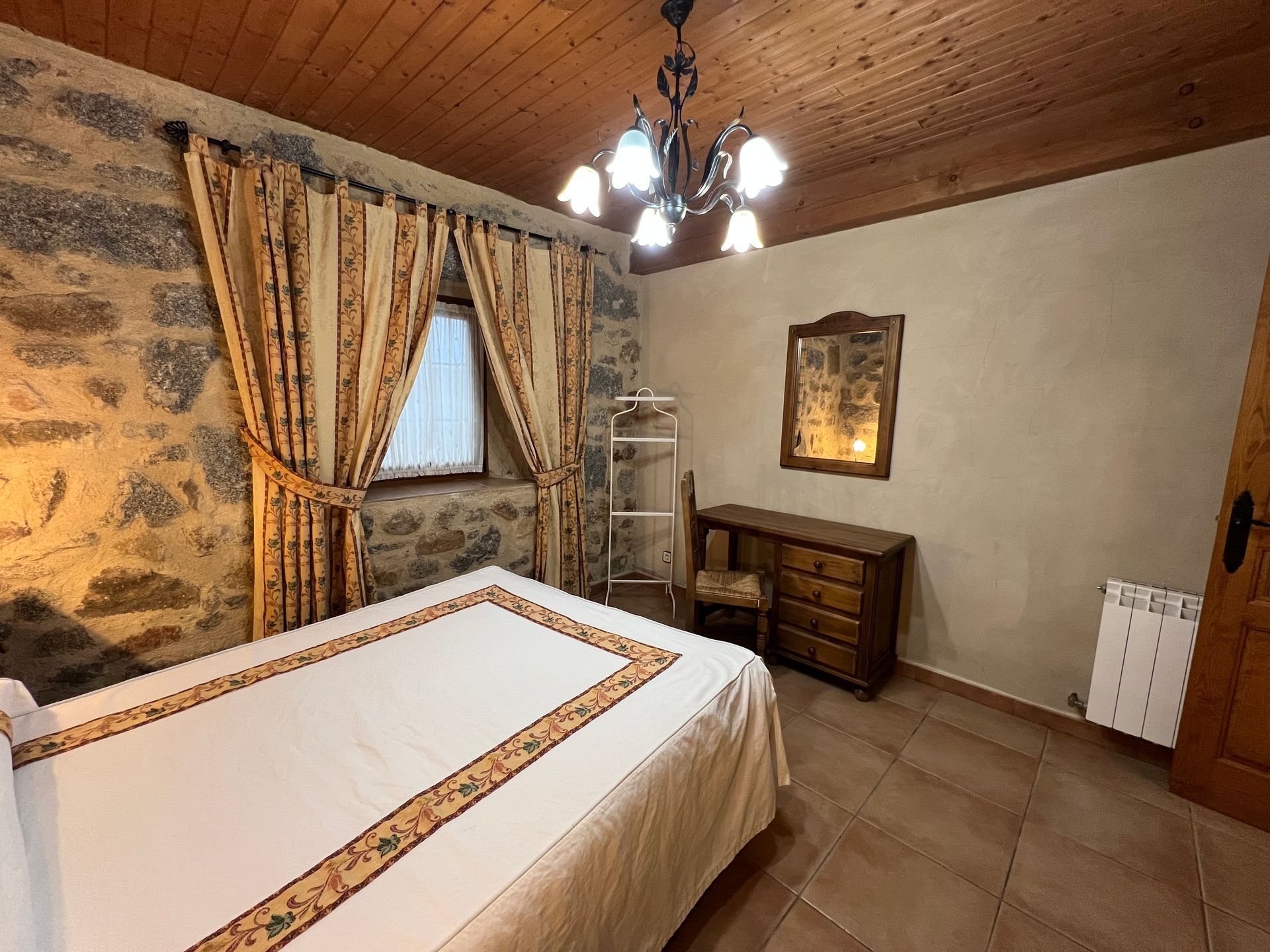 Traditional bedroom with a large bed, stone accent wall, wooden ceiling, chandelier, and dresser