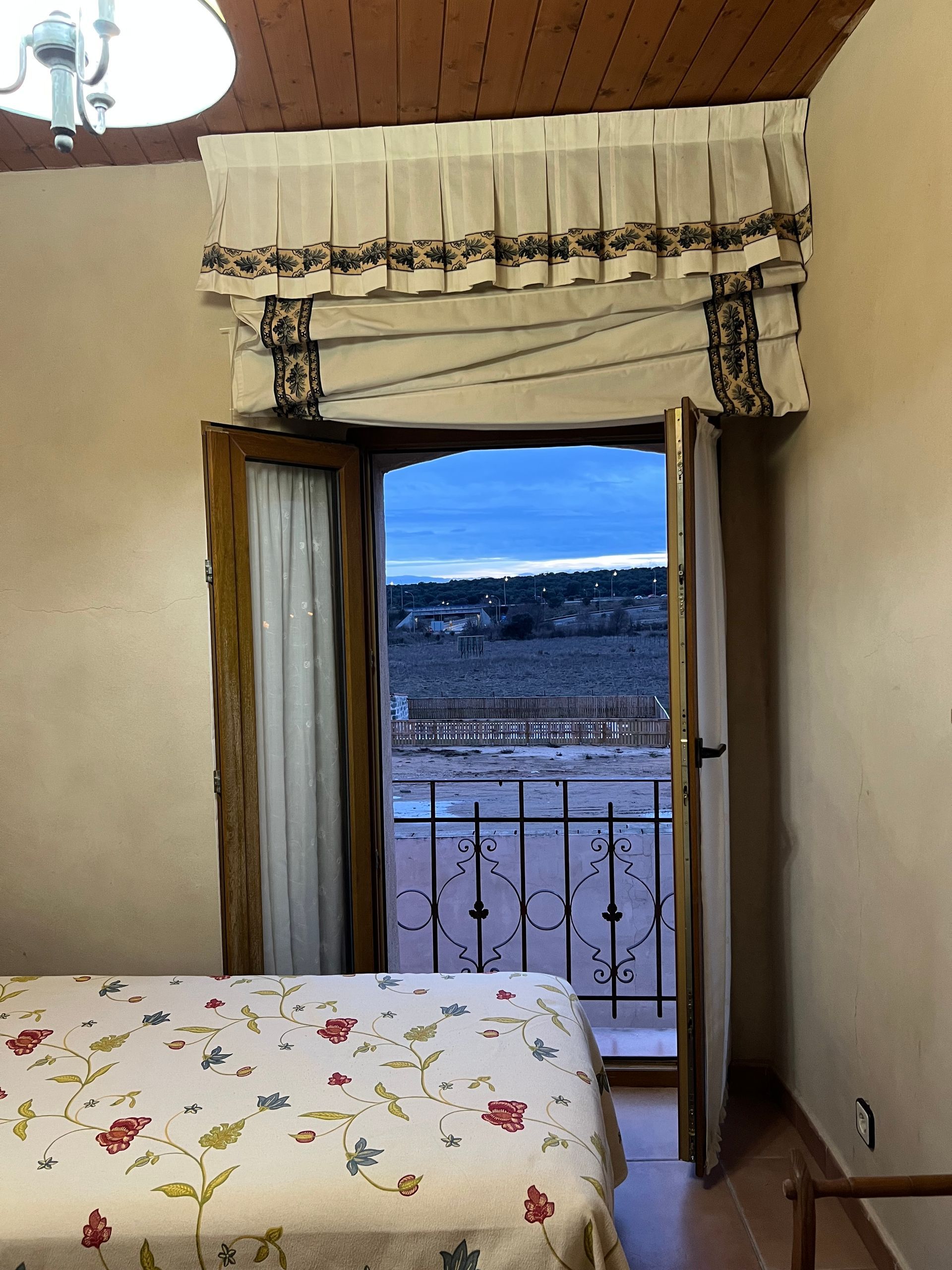 Open doorway to a balcony with railing and distant hills, seen from a bedroom with floral bedspread.