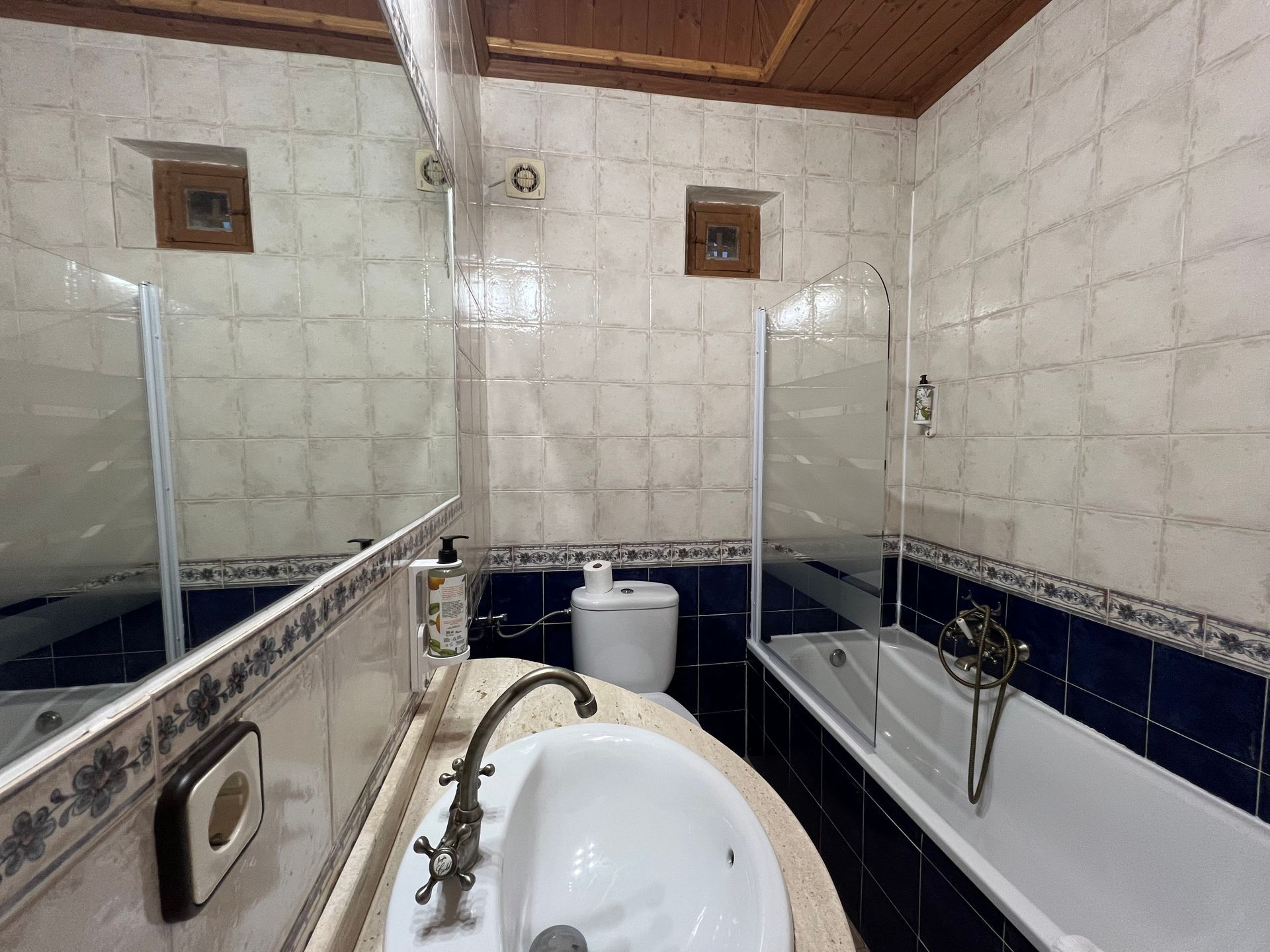 Small bathroom with two bathtubs, a central sink, mirrors, and tiled walls under a wood ceiling.