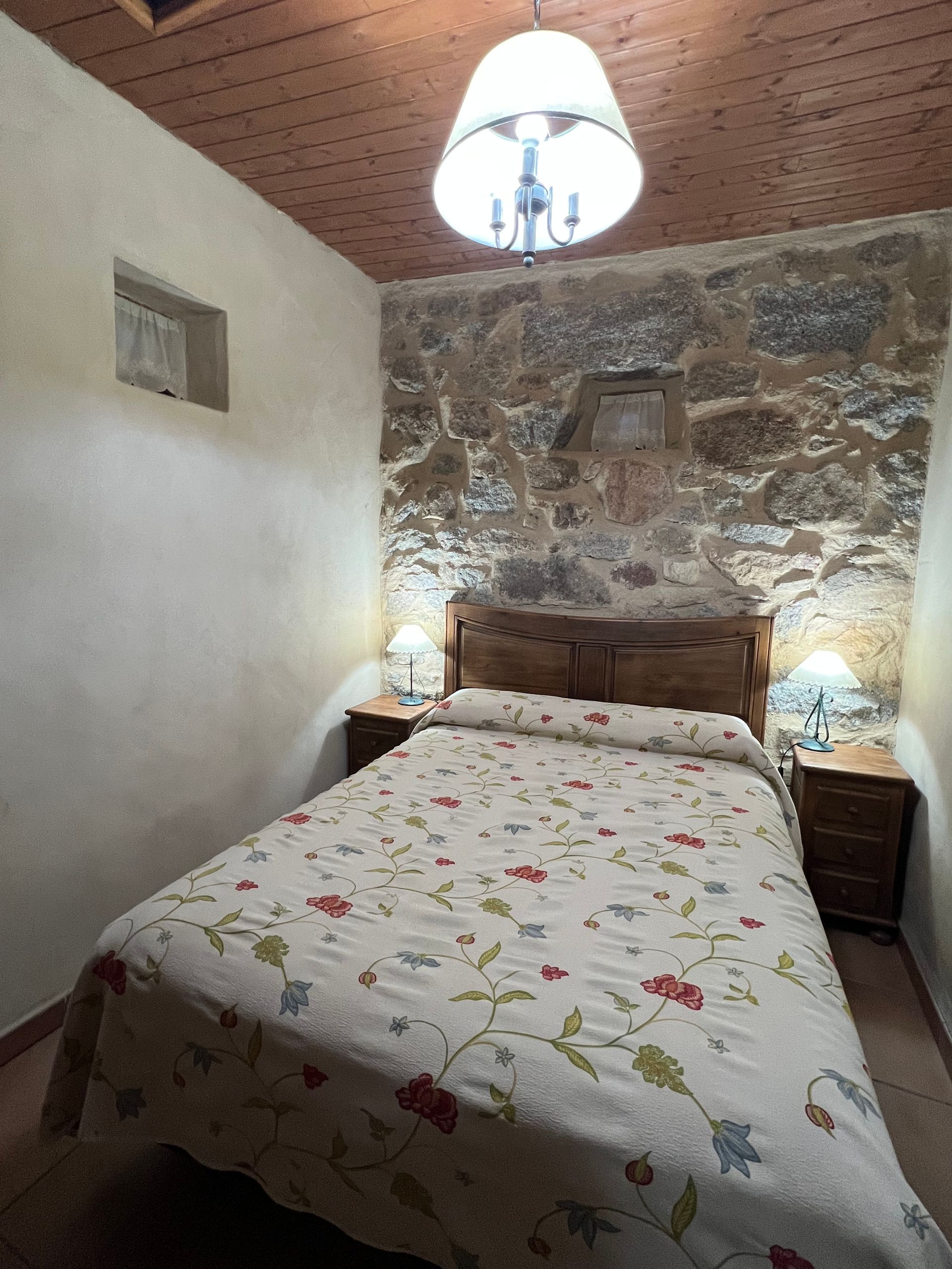 Small rustic bedroom with a floral bedspread, stone accent wall, wooden headboard, and hanging lamp.