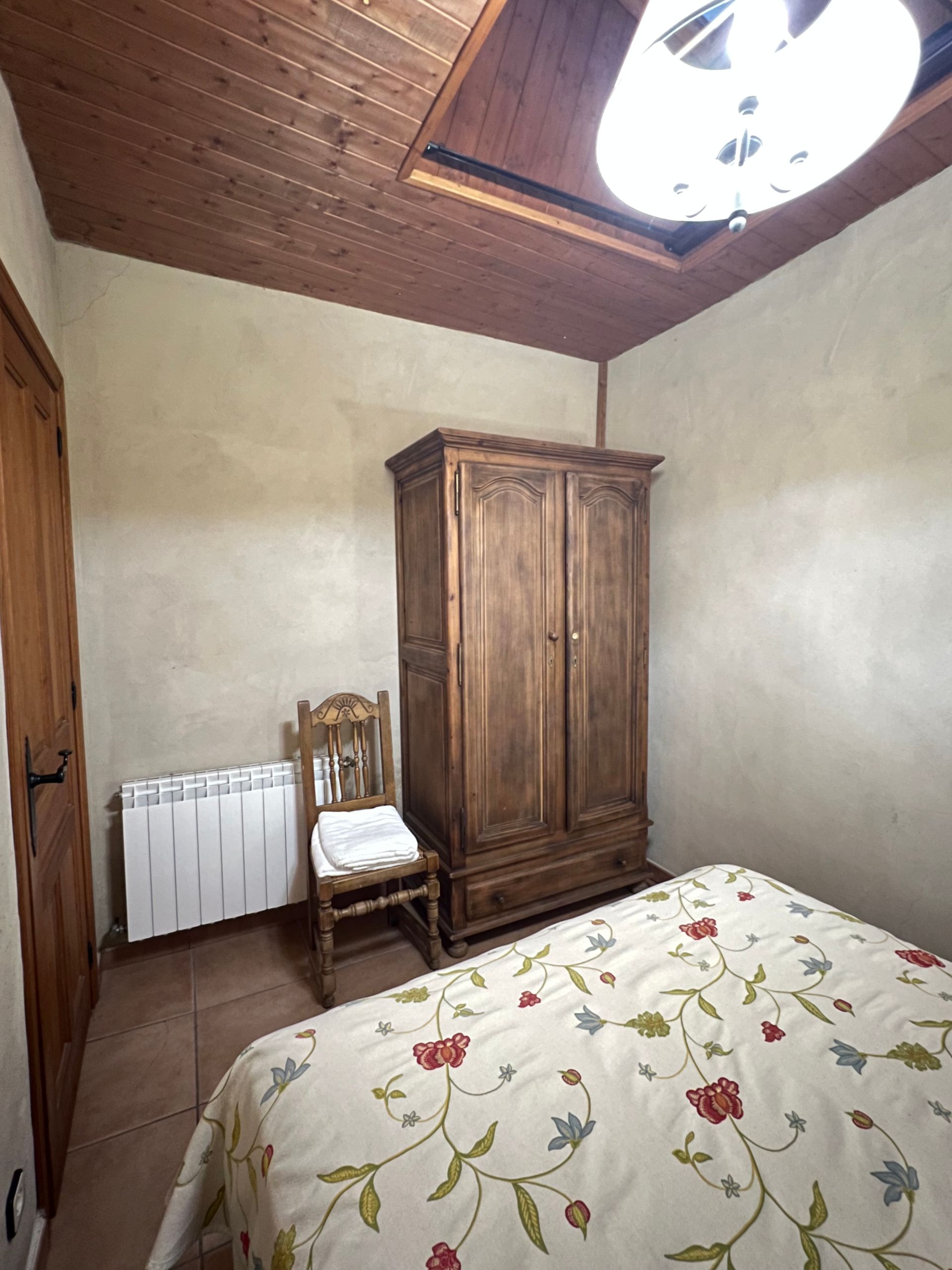 Bedroom with wooden wardrobe, chair, radiator, and floral bedspread under a wooden ceiling.