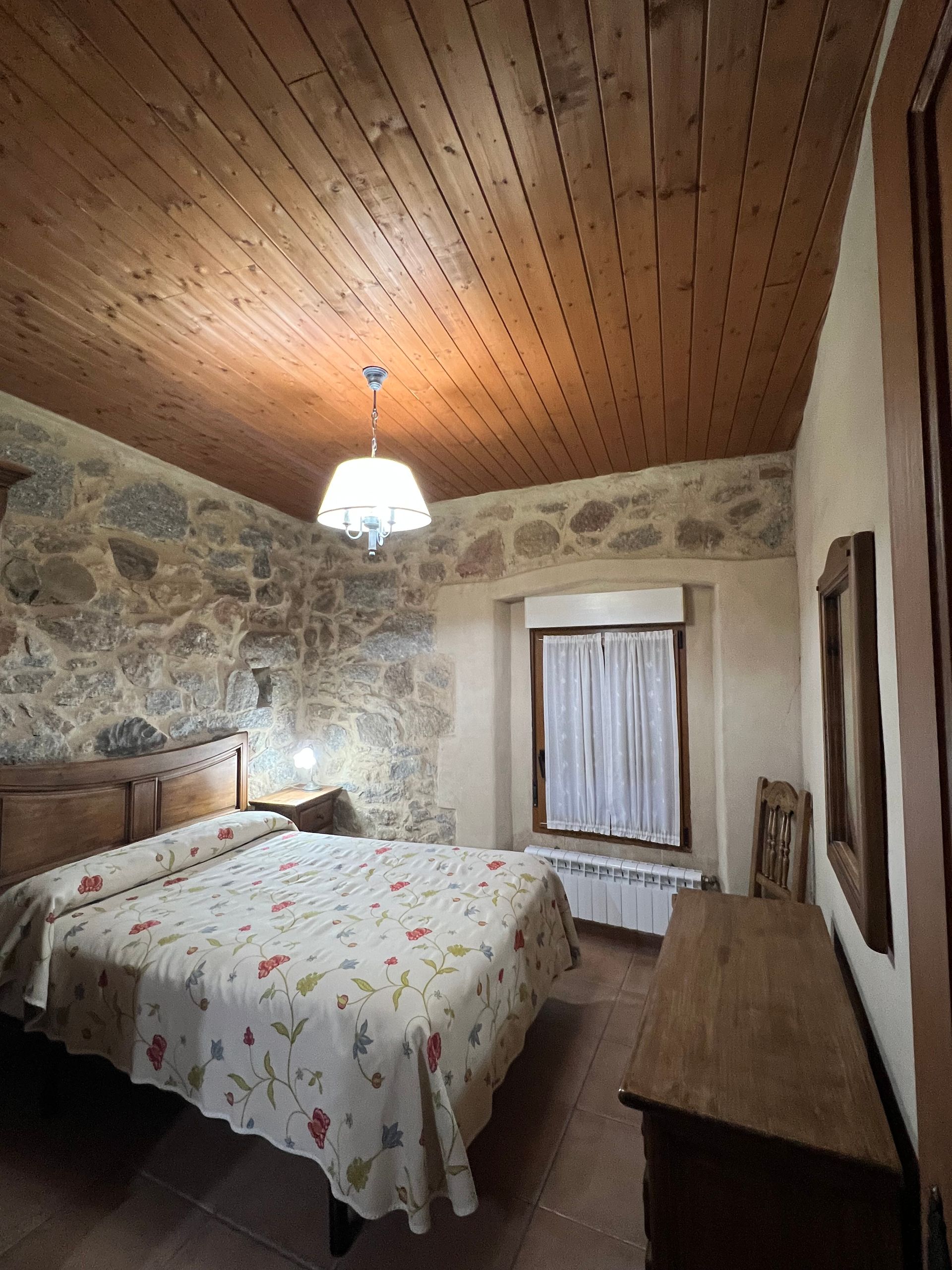 Stone-walled bedroom with wood ceiling, double bed, hanging lamp, and window with curtains