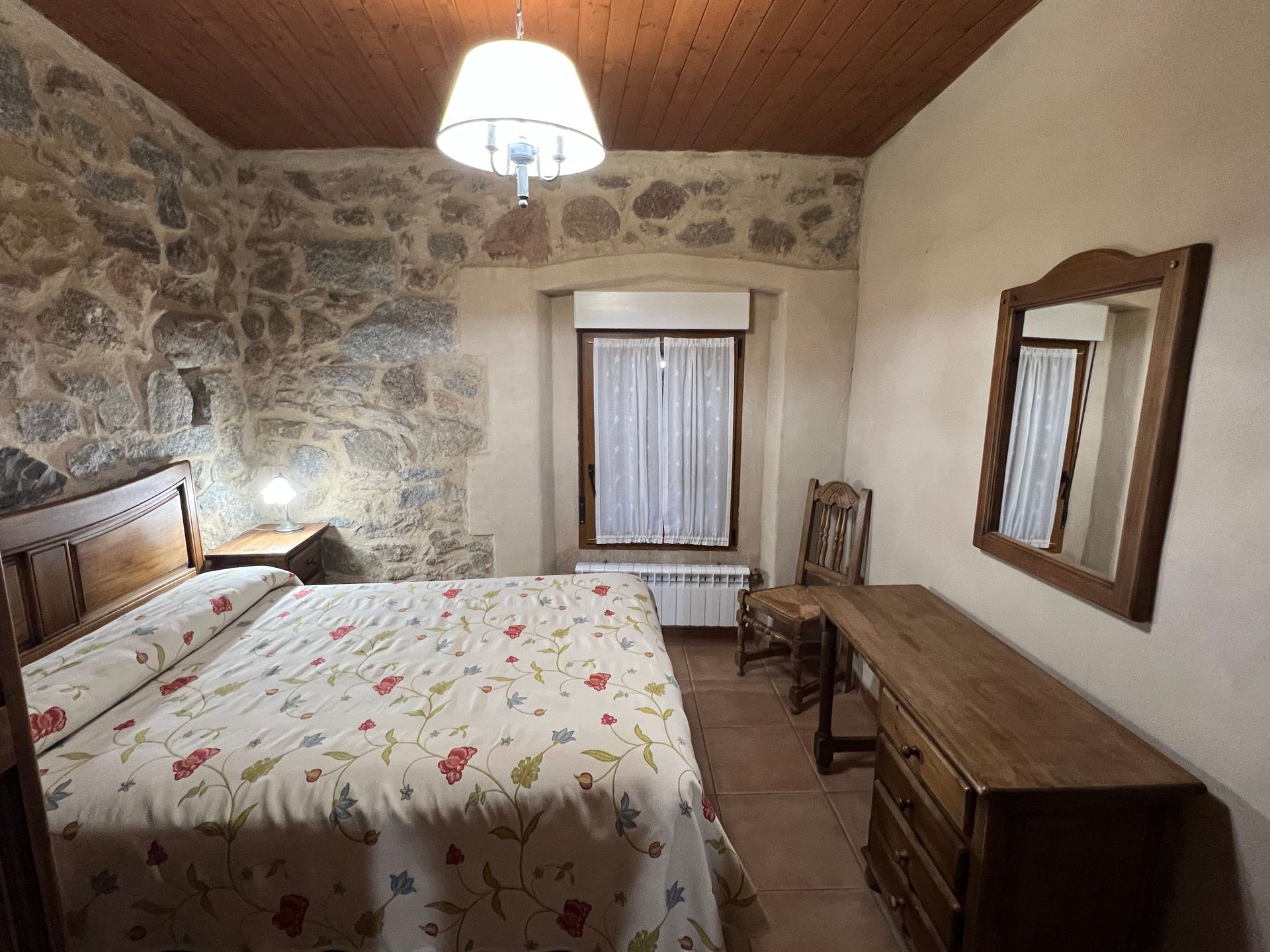 Rustic bedroom with stone walls, floral bedspread, wooden dresser, chair, and window with curtains