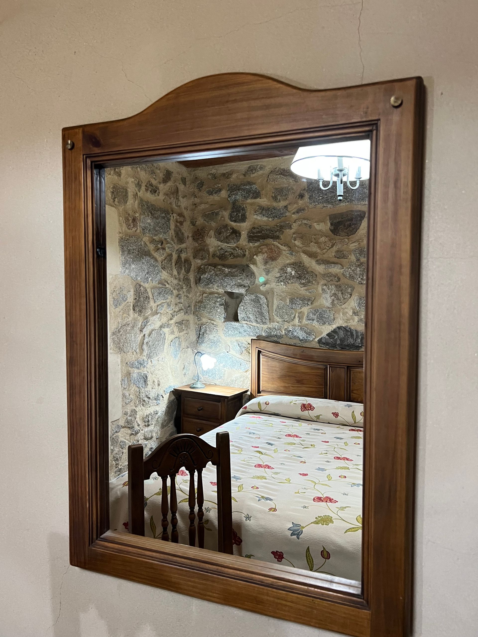 Wooden-framed mirror reflecting a bedroom with a bed, chair, patterned wallpaper, and chandelier.