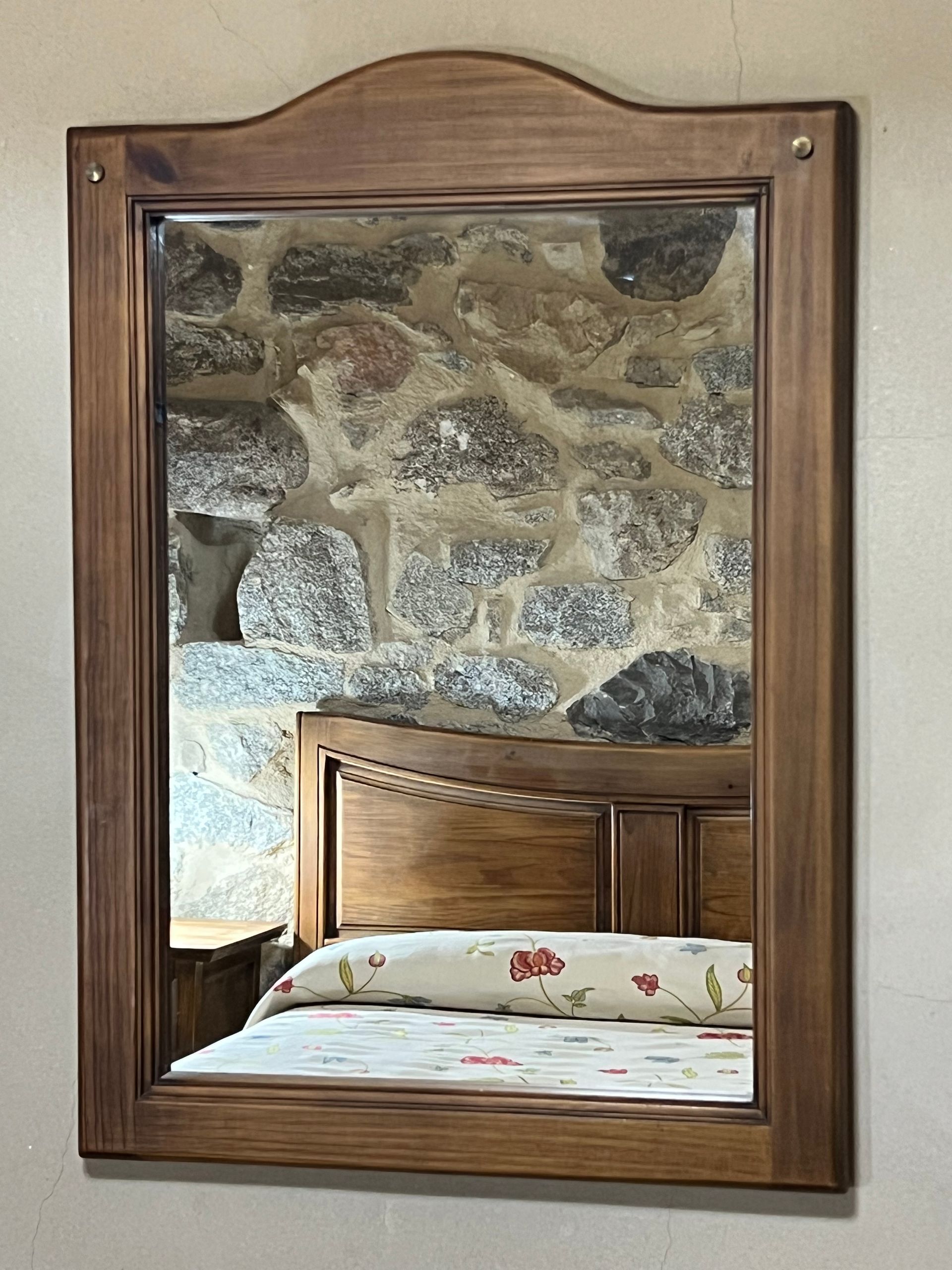 Wooden-framed mirror reflecting a bedroom headboard and patterned bedspread against a stone wall.