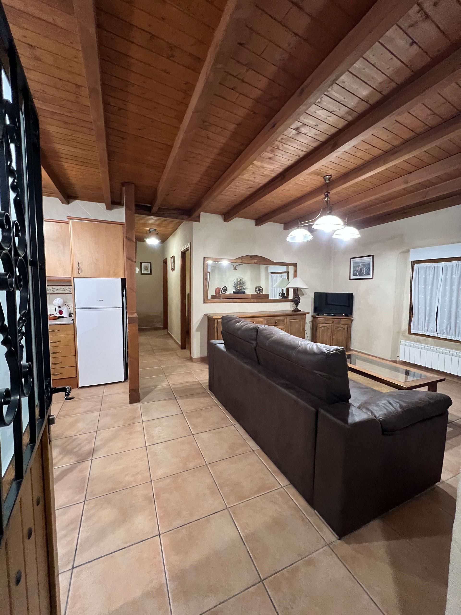 Rustic open-plan living room with wood-beamed ceiling, tiled floor, dark sofa, fridge, and kitchen area.