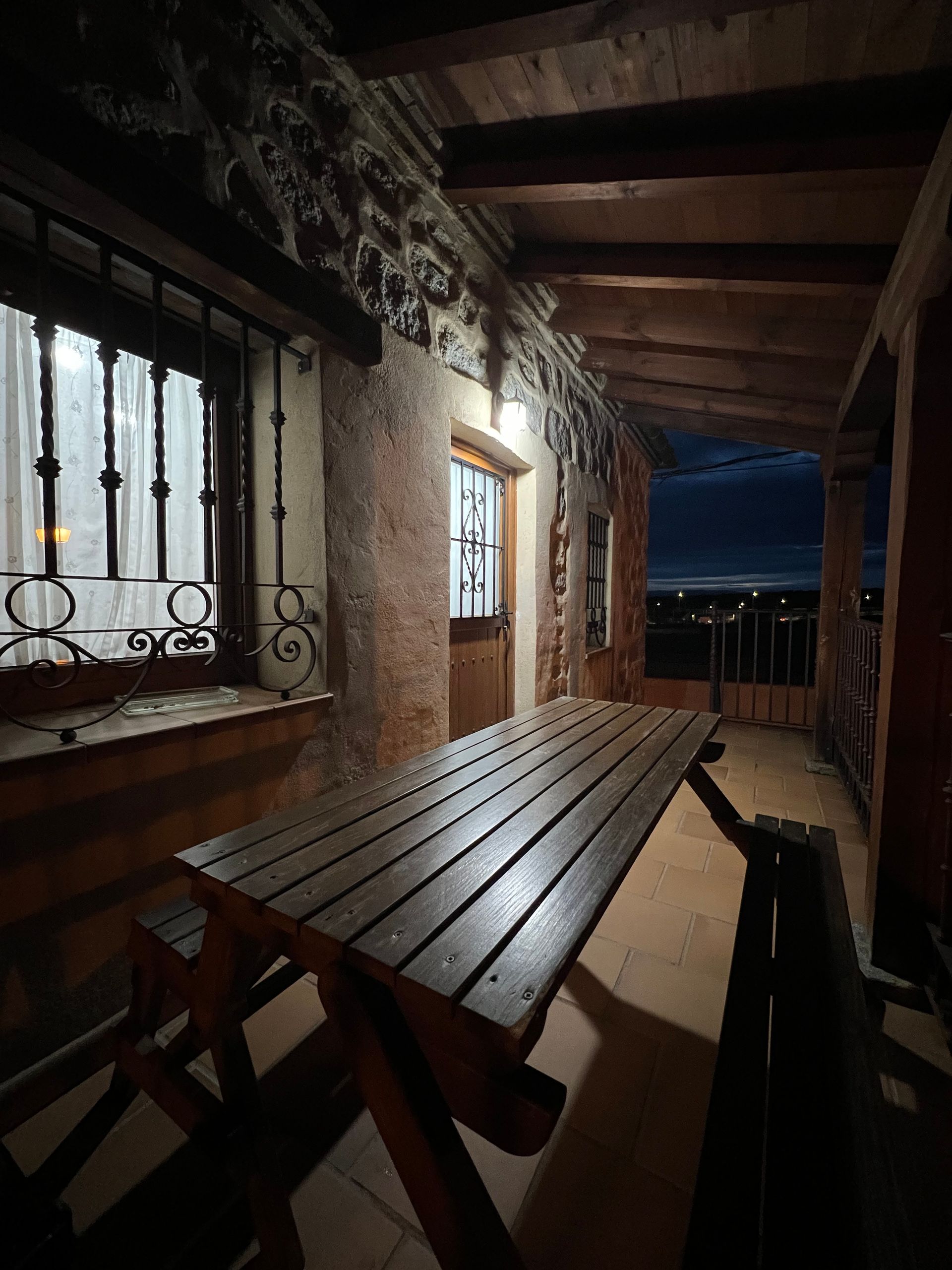 Rustic covered porch with long wooden picnic table, stone wall, and dark evening view beyond the railing