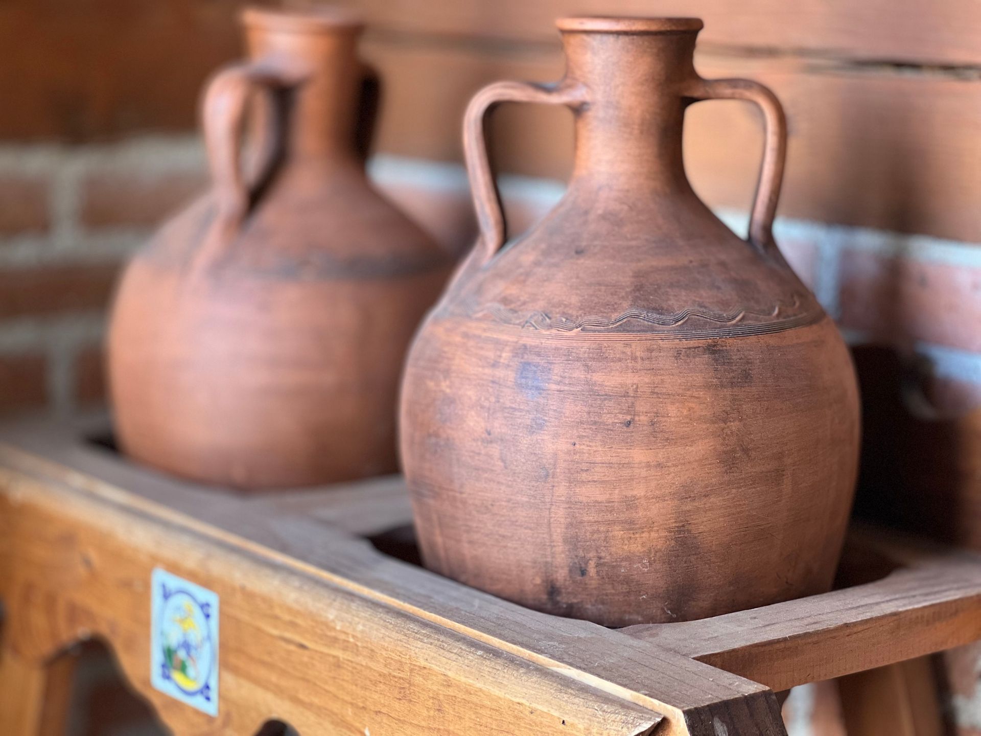 Two terracotta clay jugs with handles in a wooden display tray, against a brick wall.