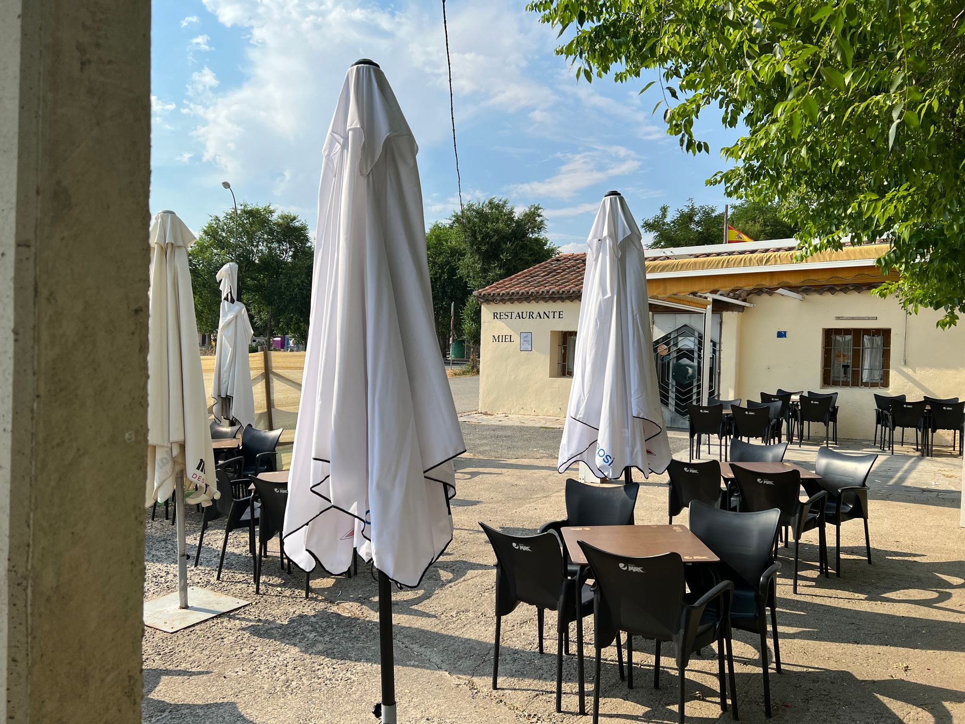 Outdoor café terrace with closed white umbrellas, black chairs, and stone tables under trees and blue sky