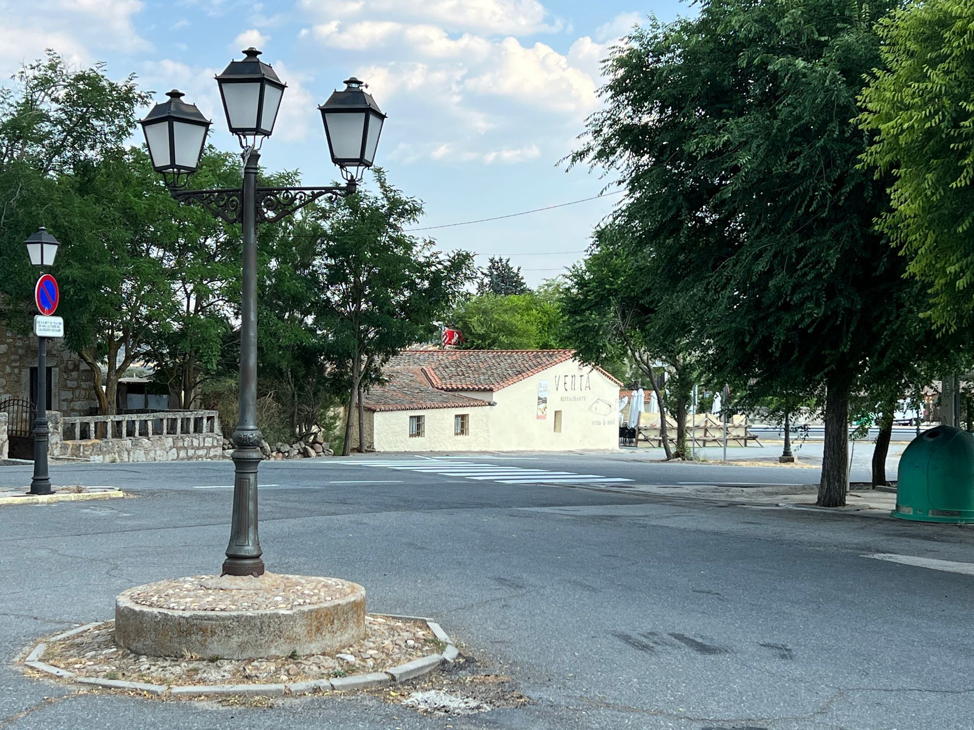 Quiet cobblestone square with a three-lantern streetlight, trees, and a pale building in the background