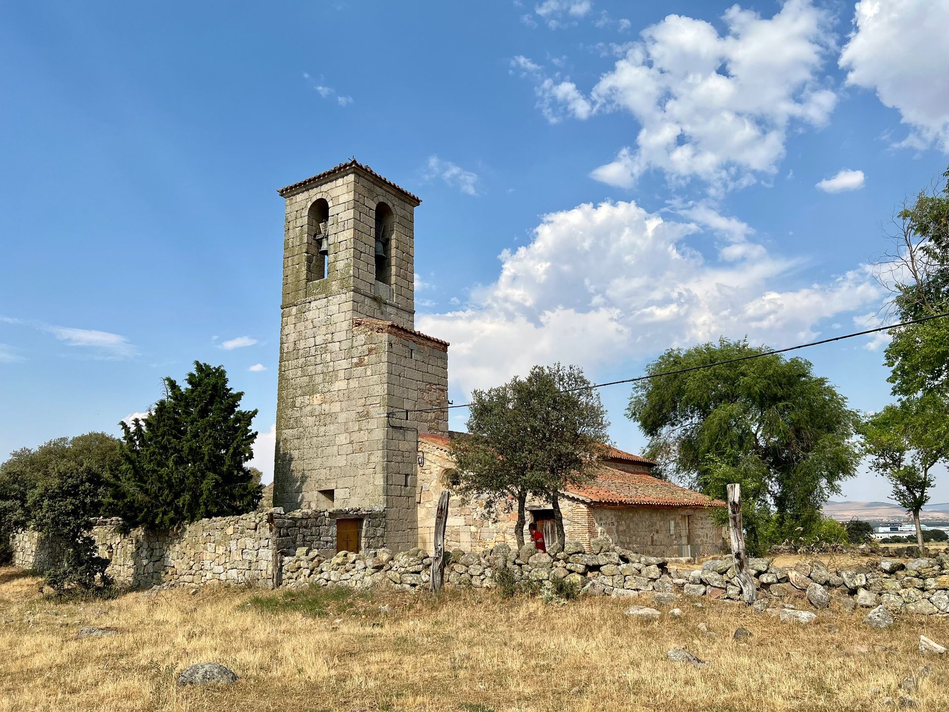 Stone church tower and small building in a dry field under a blue sky with scattered clouds