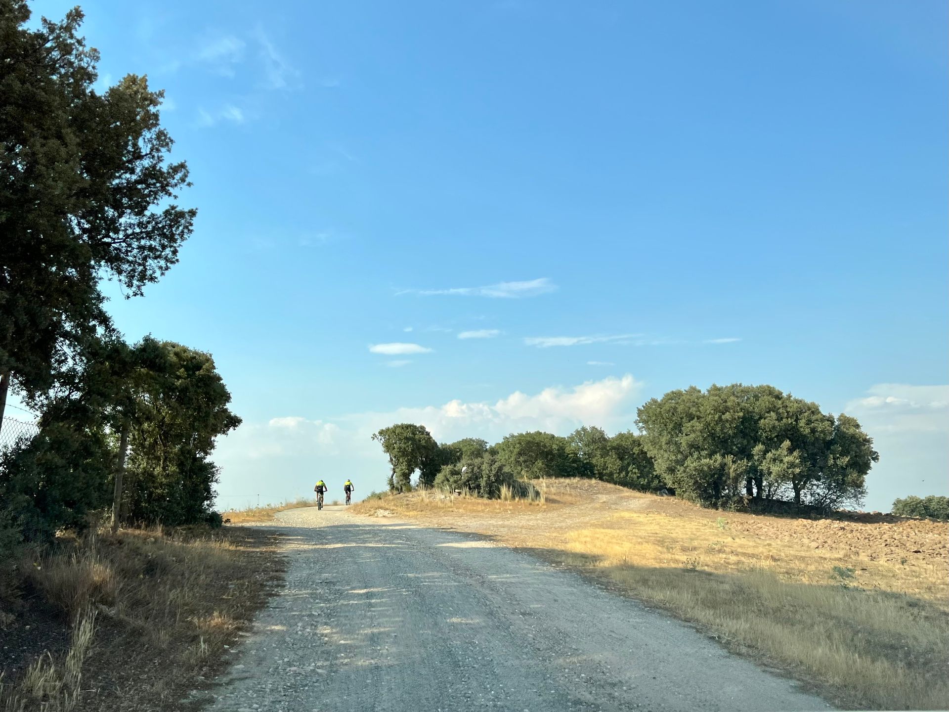 Gravel road rising toward a sunlit hill with trees and two small people in the distance under a clear blue sky