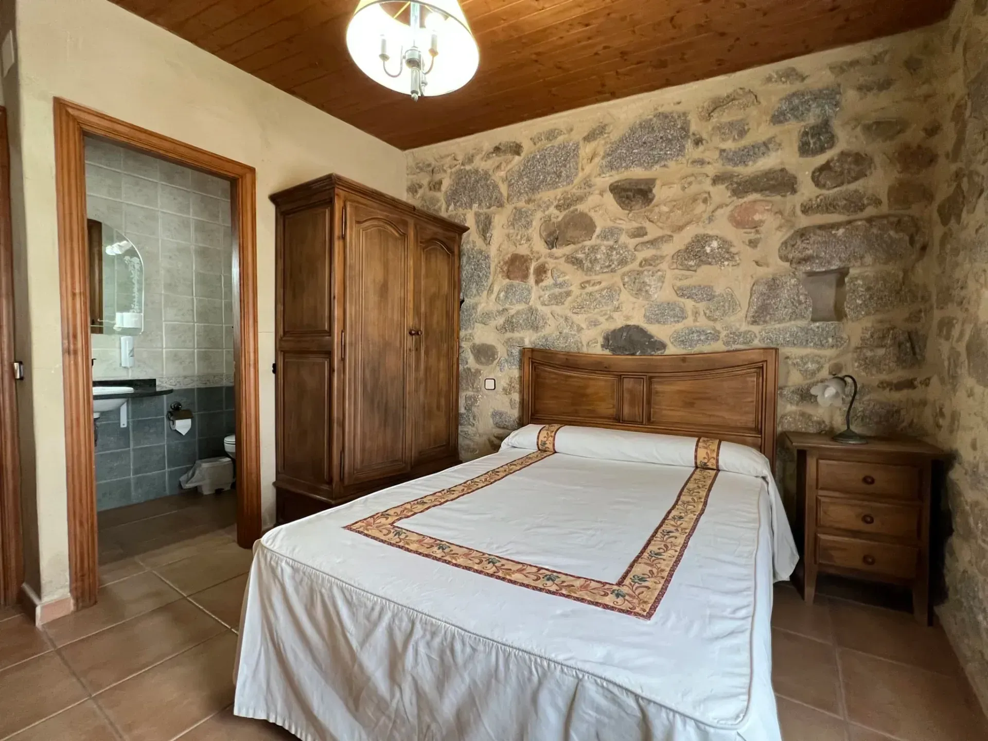 Rustic bedroom with wooden bed, stone wall, wardrobe, nightstand, and tiled floor
