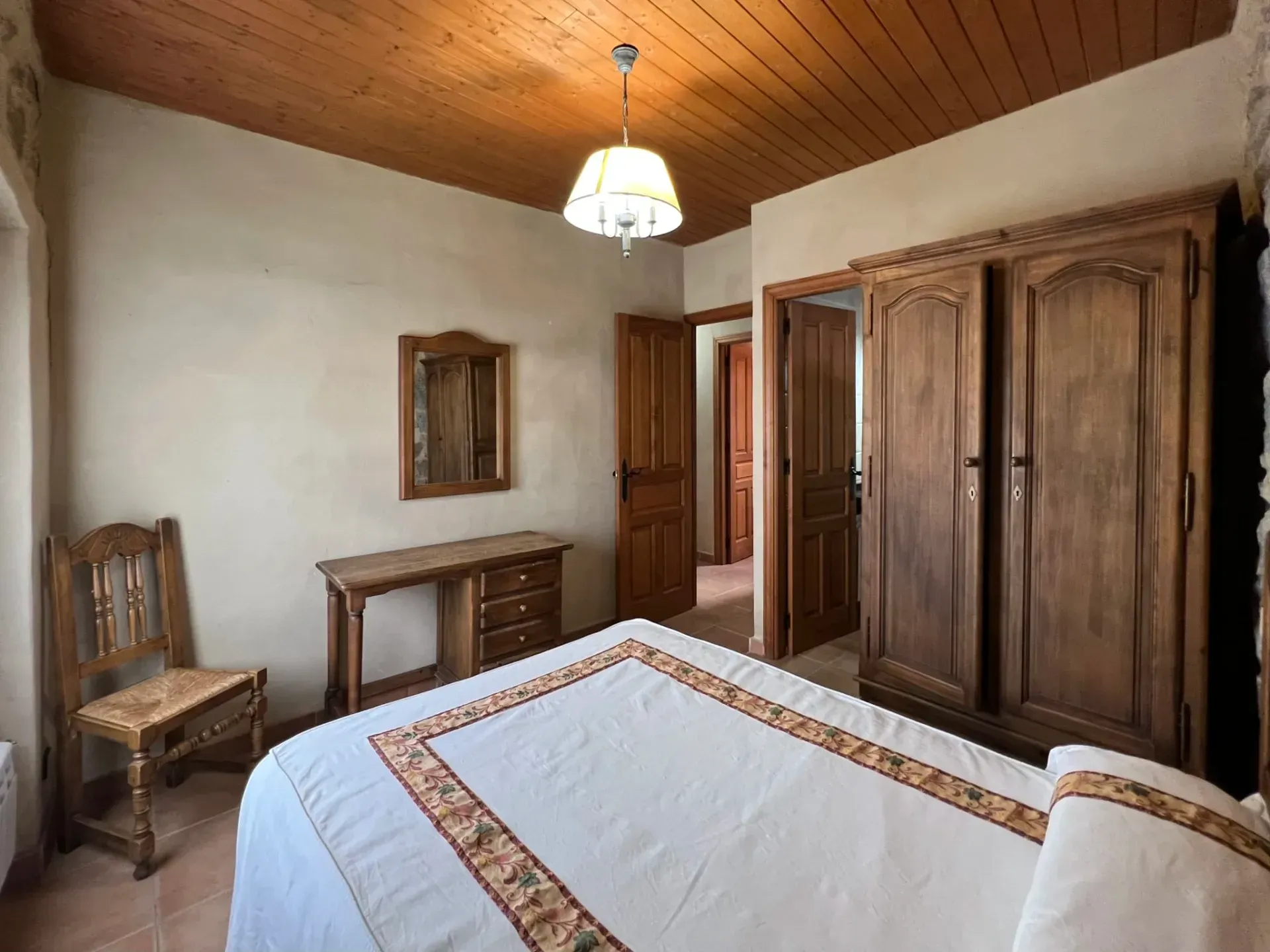 Rustic bedroom with wooden ceiling, large wardrobe, desk, chair, and bed with patterned bedspread