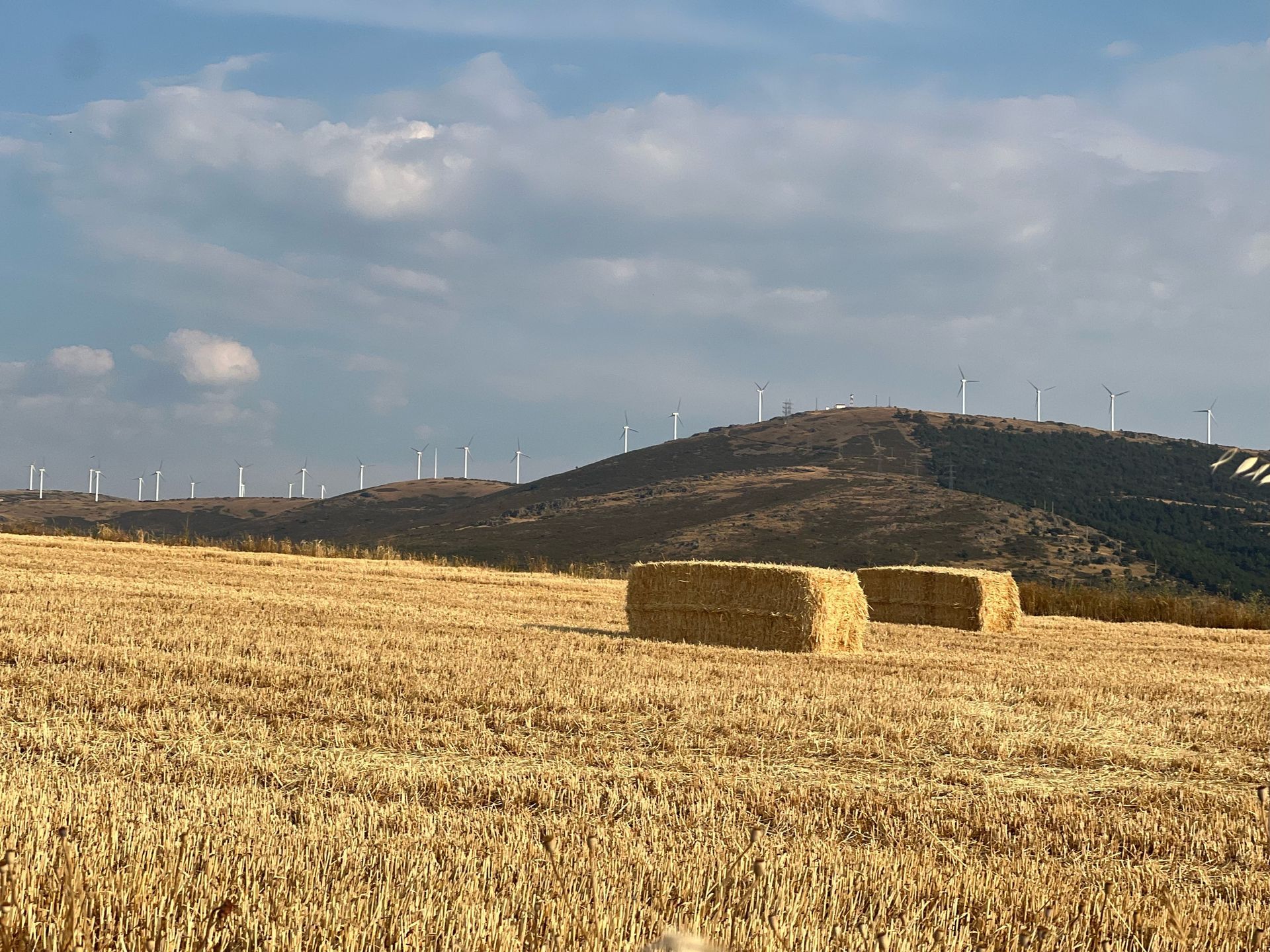 Hay bales in a golden field with wind turbines on a distant hill under a partly cloudy sky