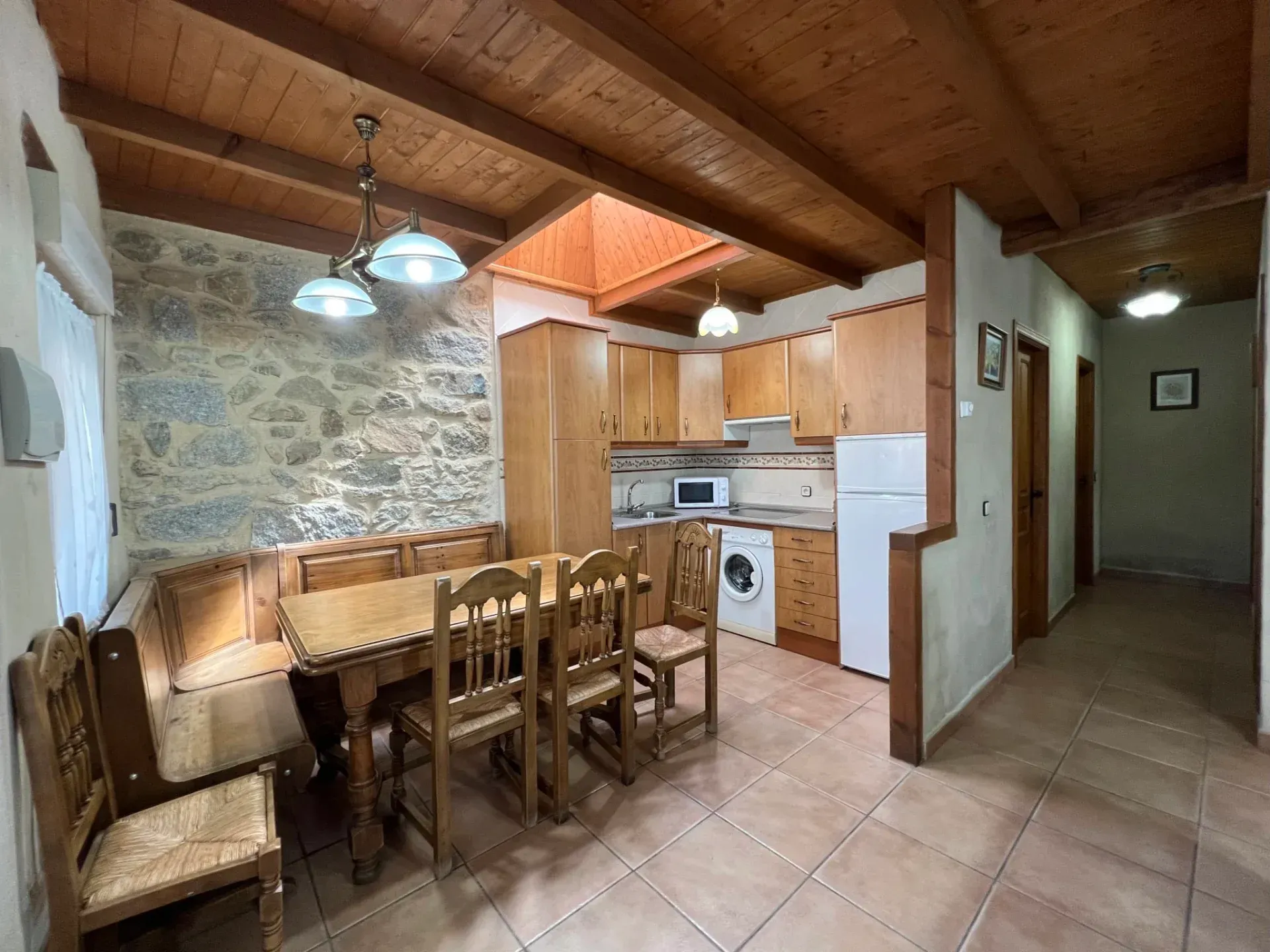 Rustic dining area with wooden table, chairs, stone wall, and compact kitchen under sloped wood ceiling