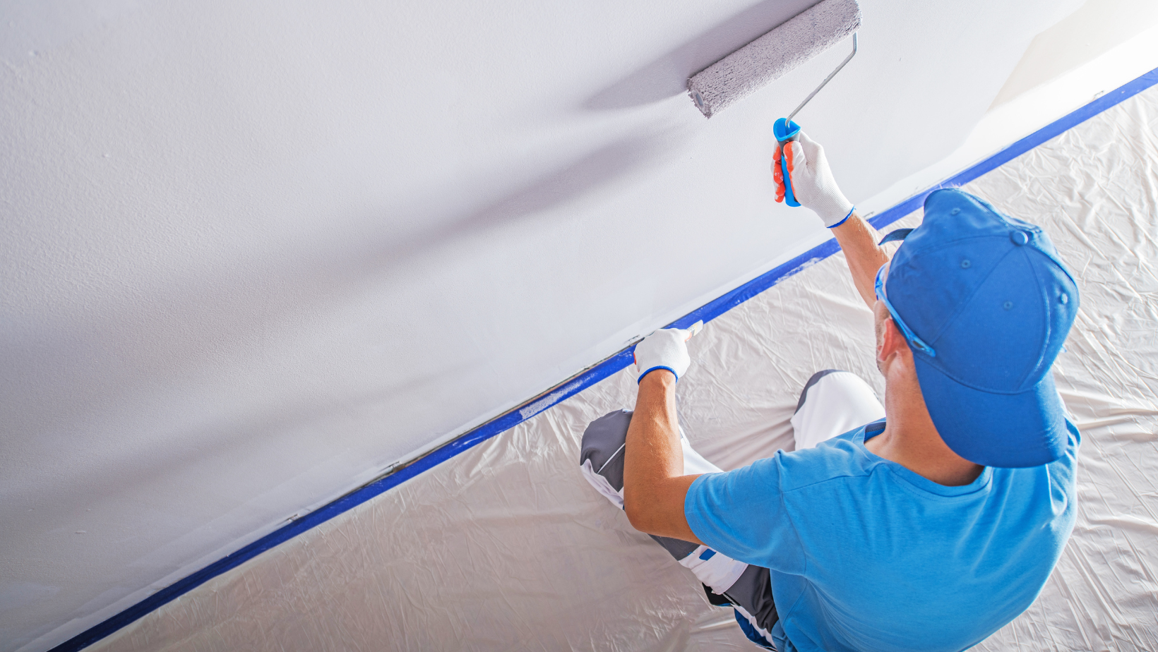 Person painting a ceiling with a paint roller, wearing blue work clothes and a cap.