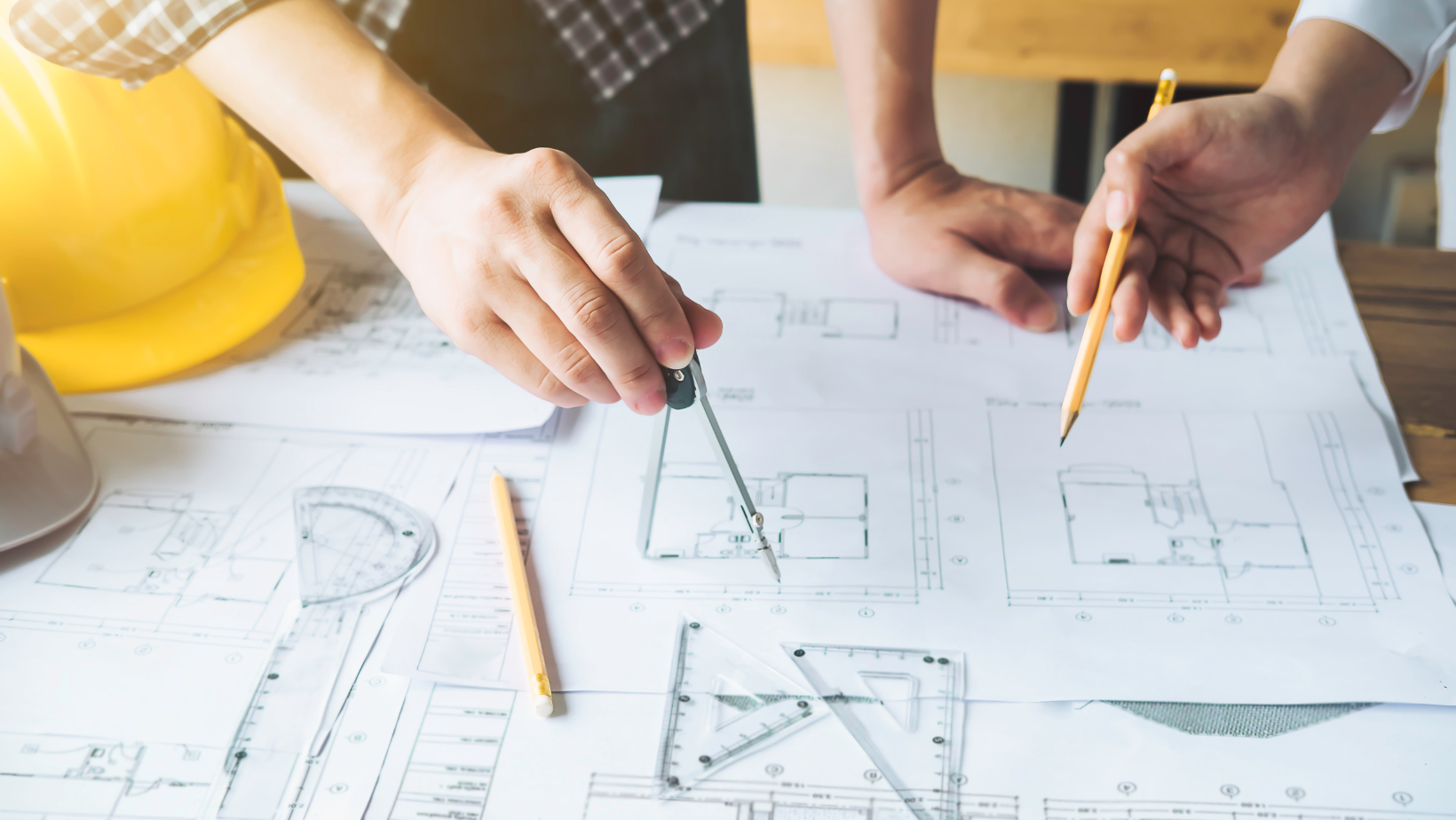 Hands using compass and pencil on architectural blueprints, yellow hard hat nearby.