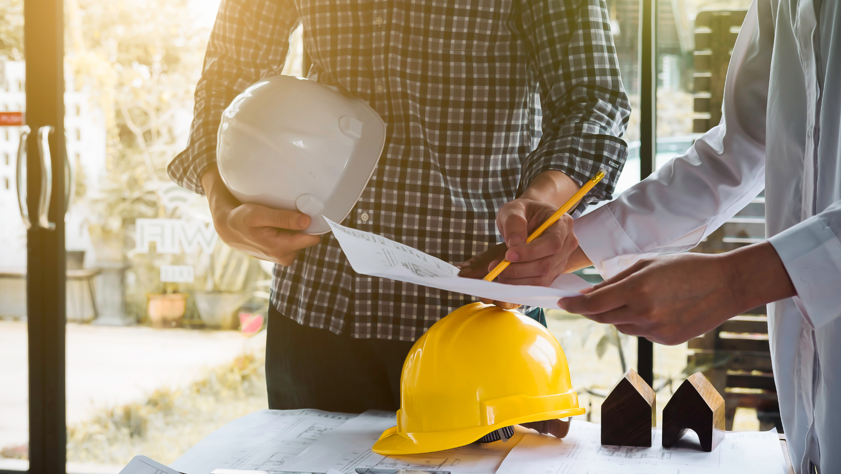 Two people reviewing blueprints, one holding a white hard hat, yellow hard hat on table.