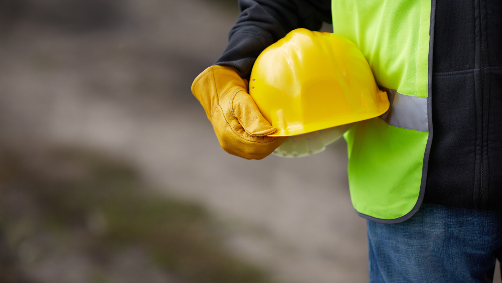 Person in a neon safety vest holding a yellow hard hat outdoors