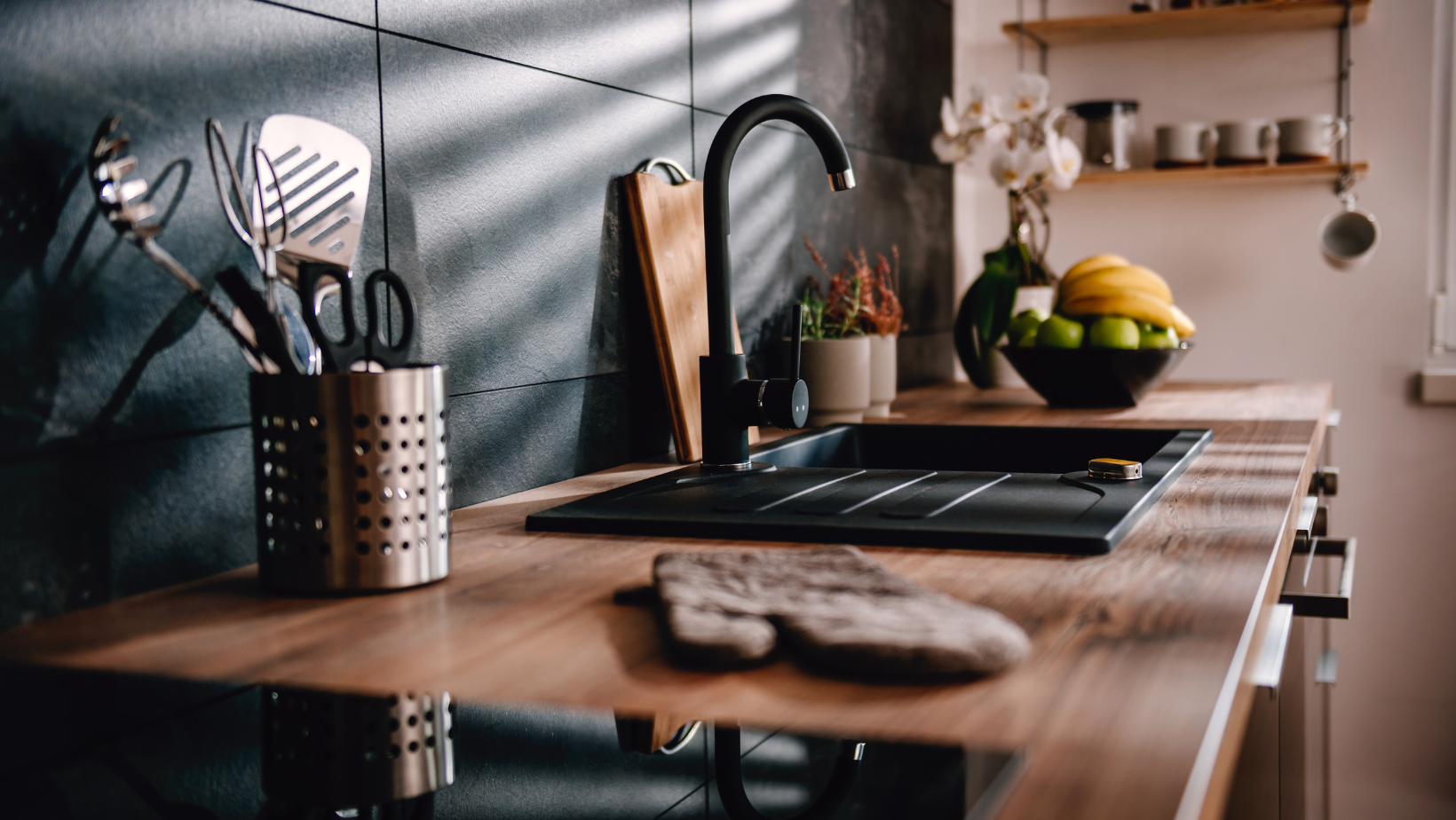Warm kitchen counter with wooden cutting board, utensil holder, stovetop, fruit bowl, and wall shelves