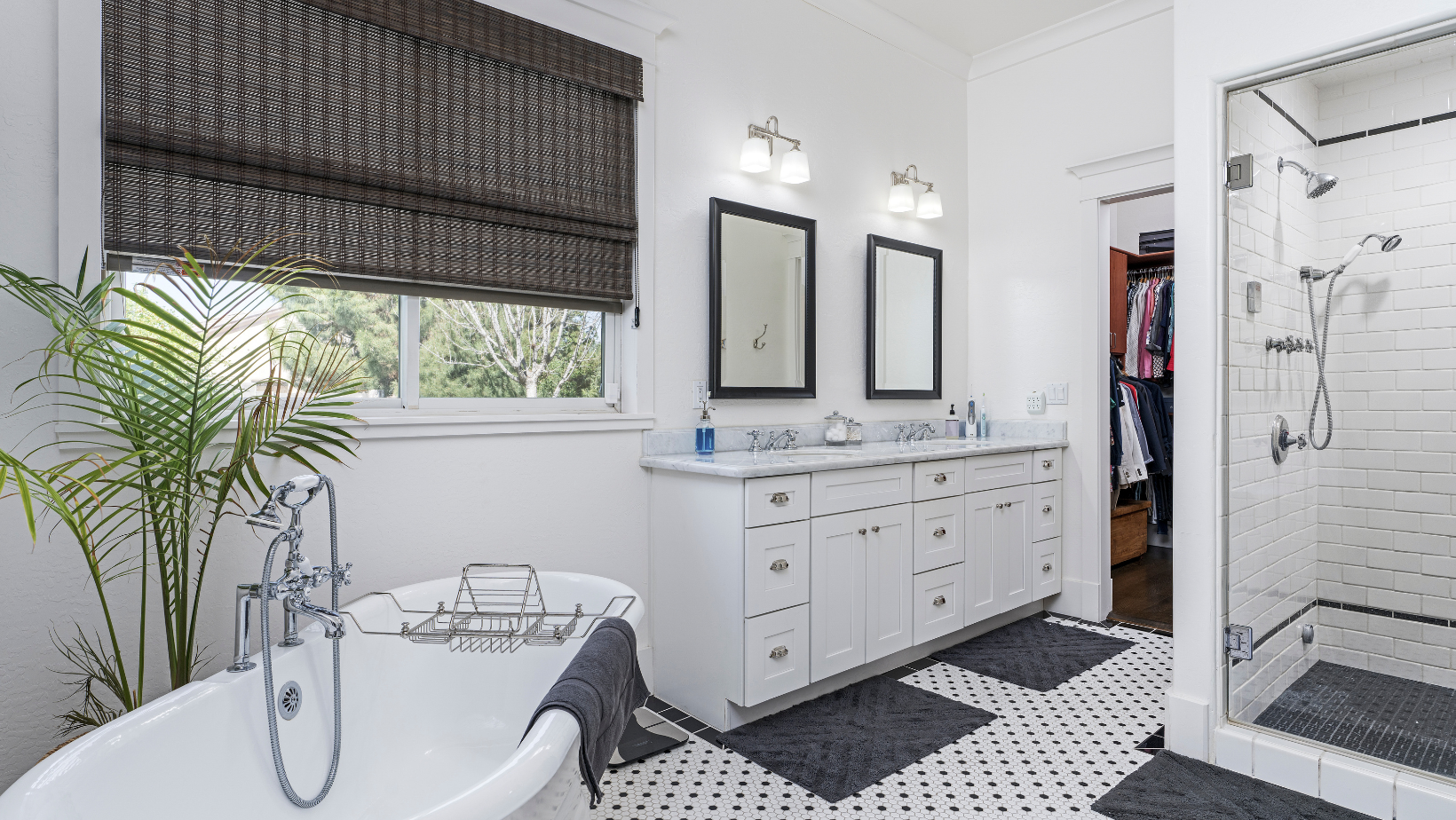 Bright bathroom with freestanding tub, white vanity, large shower, black-and-white floor tiles, and a window with blinds