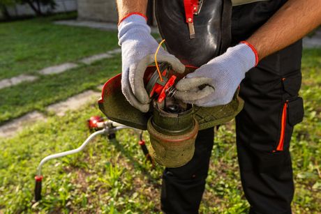 A person wearing work gloves repairs the spool of a string trimmer outdoors on a lawn.