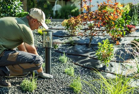 A person wearing a cap and work clothes kneels in a garden to install an outdoor light fixture in a gravel bed.