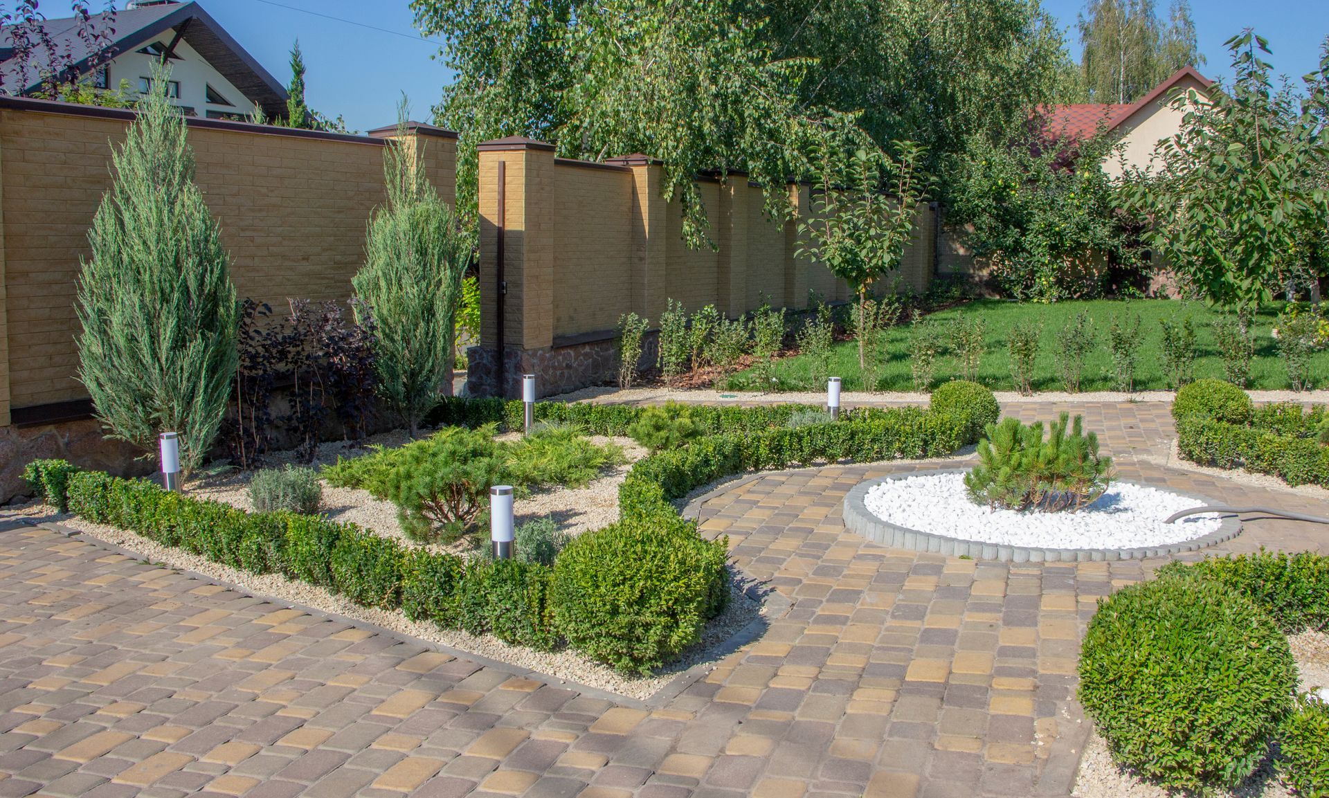 A modern garden with manicured hedges, shaped bushes, conifer trees, and paved paths against a brick wall.