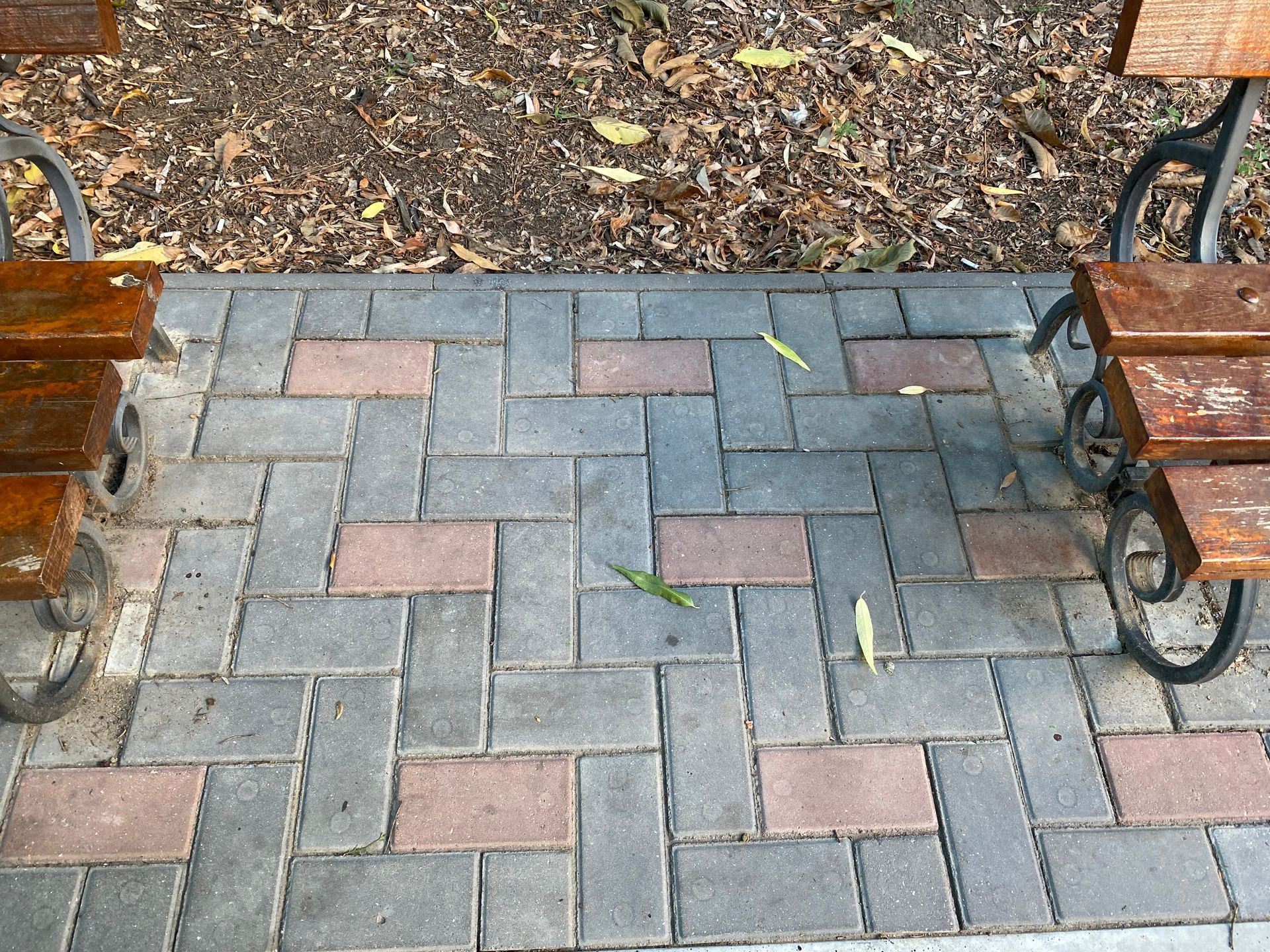 Two wooden park benches sit on either side of a walkway paved with a grey and red interlocking brick pattern.
