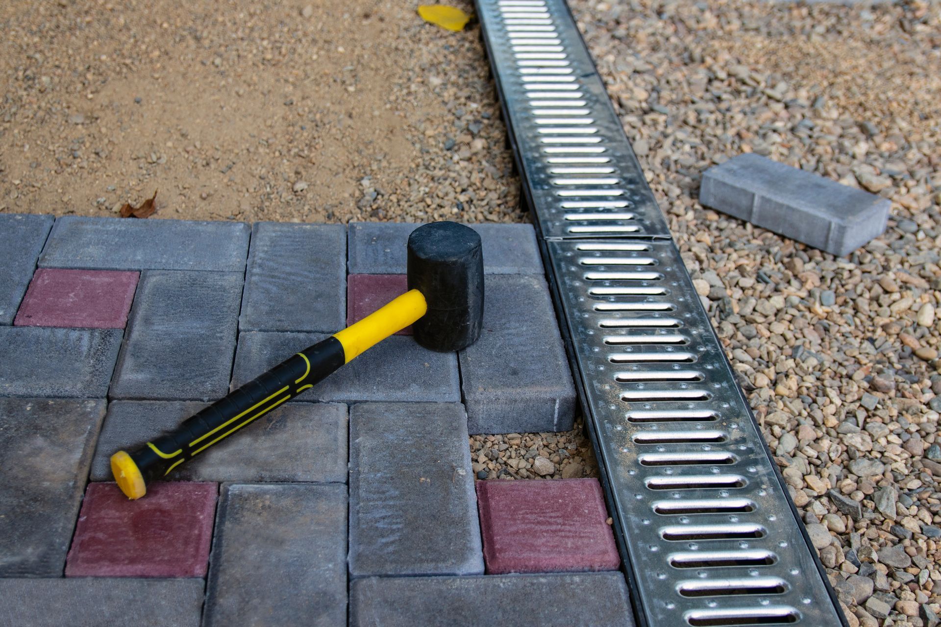 A black rubber mallet resting on gray and red paving stones next to a metal drainage grate.