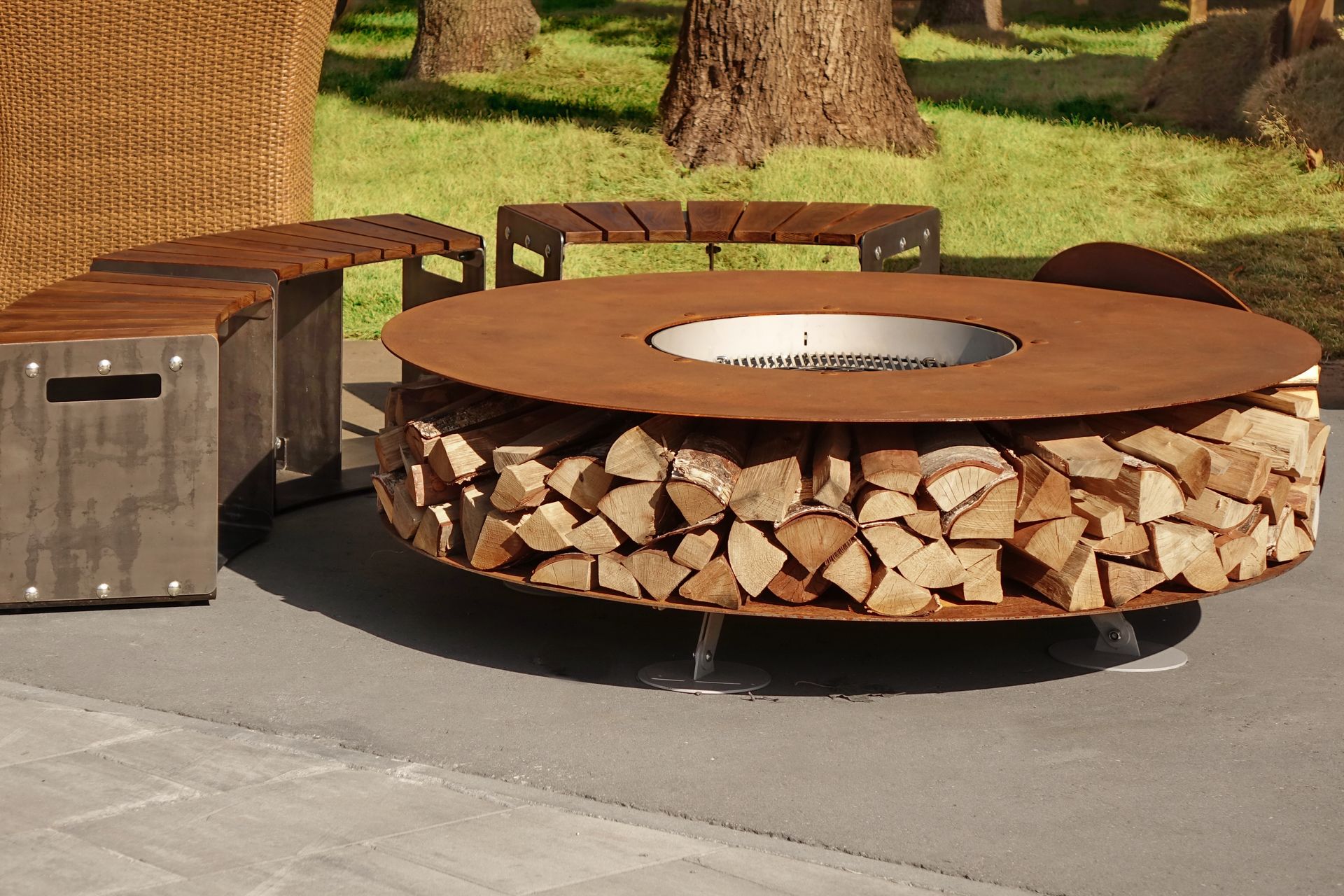 A circular, rustic metal fire pit table with a base stacked with firewood, set on a patio with matching benches.