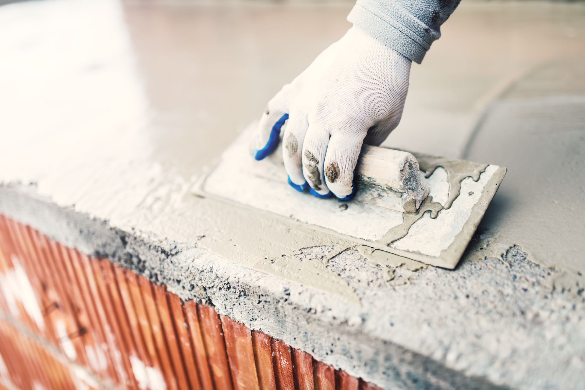 A gloved hand uses a metal trowel to smooth wet concrete onto a red brick structure.