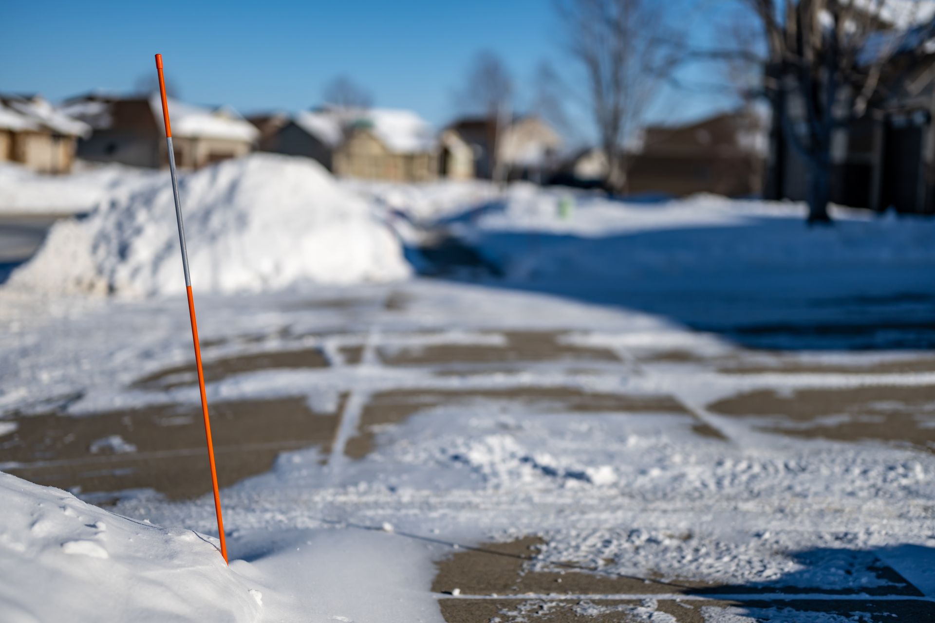 An orange reflective driveway marker stands in snow-covered pavement on a sunny day in a suburban neighborhood.