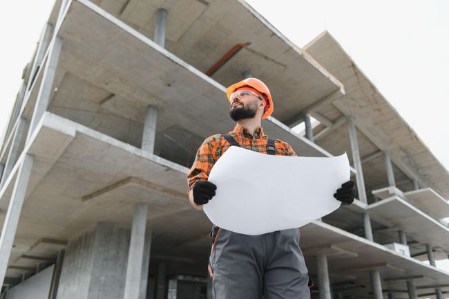 A construction worker in a hard hat and work clothes holds a blueprint in front of an unfinished concrete building.