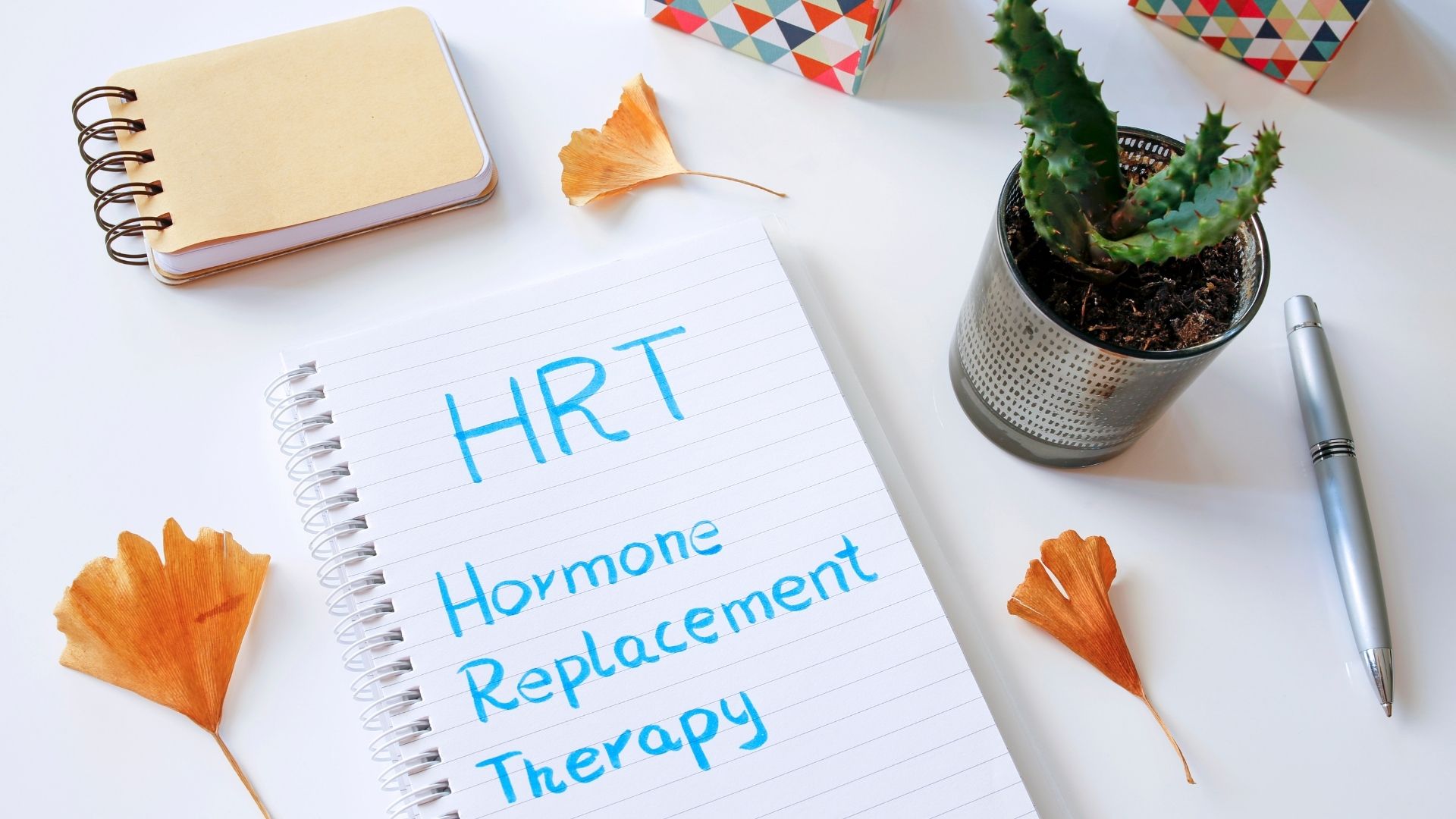 Notebook reading “HRT Hormone Replacement Therapy” on a white desk with pen, plant, and paper cranes