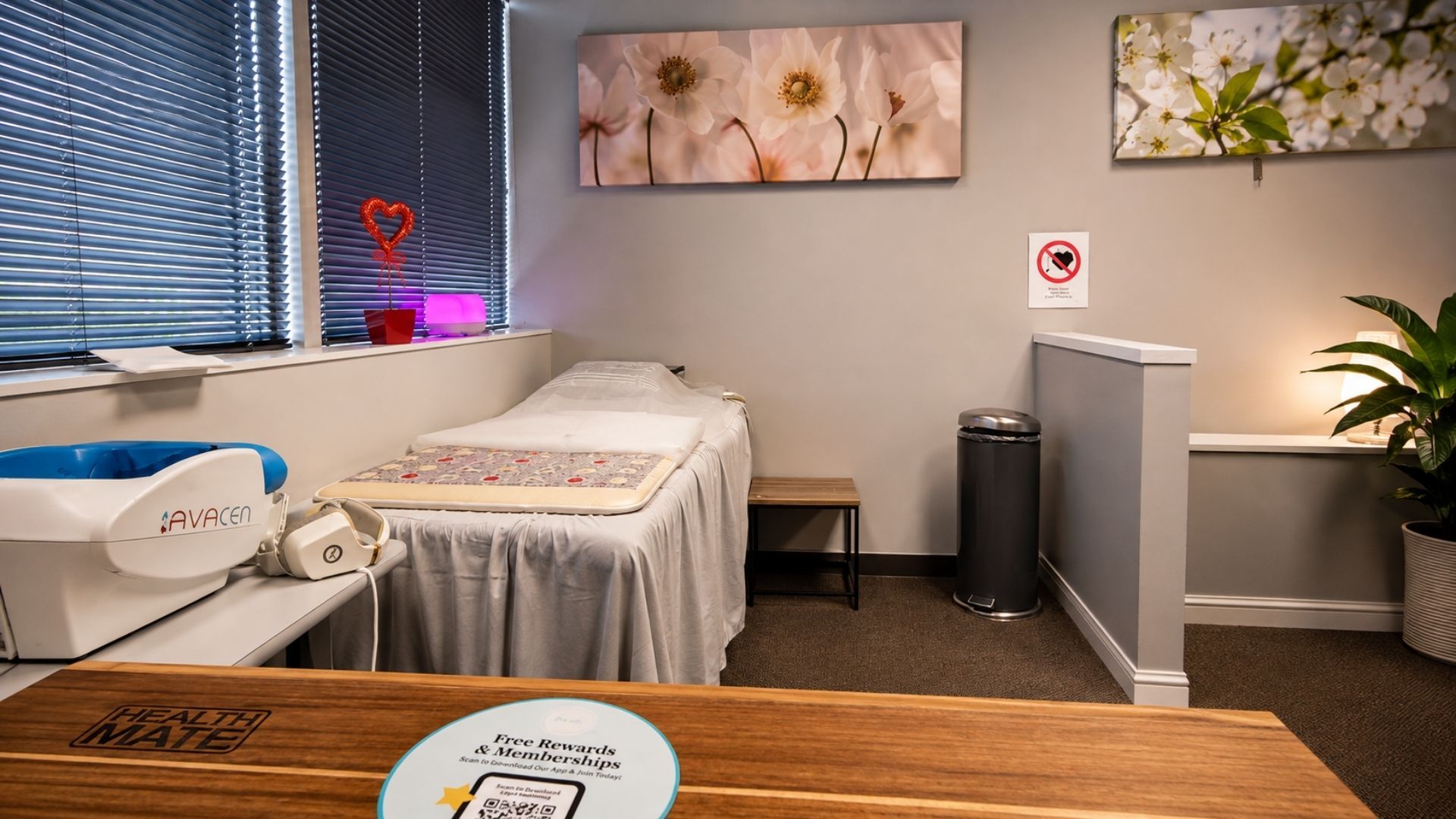 Small spa room with massage table, white chairs, floral wall art, and a desk by a window with blinds.