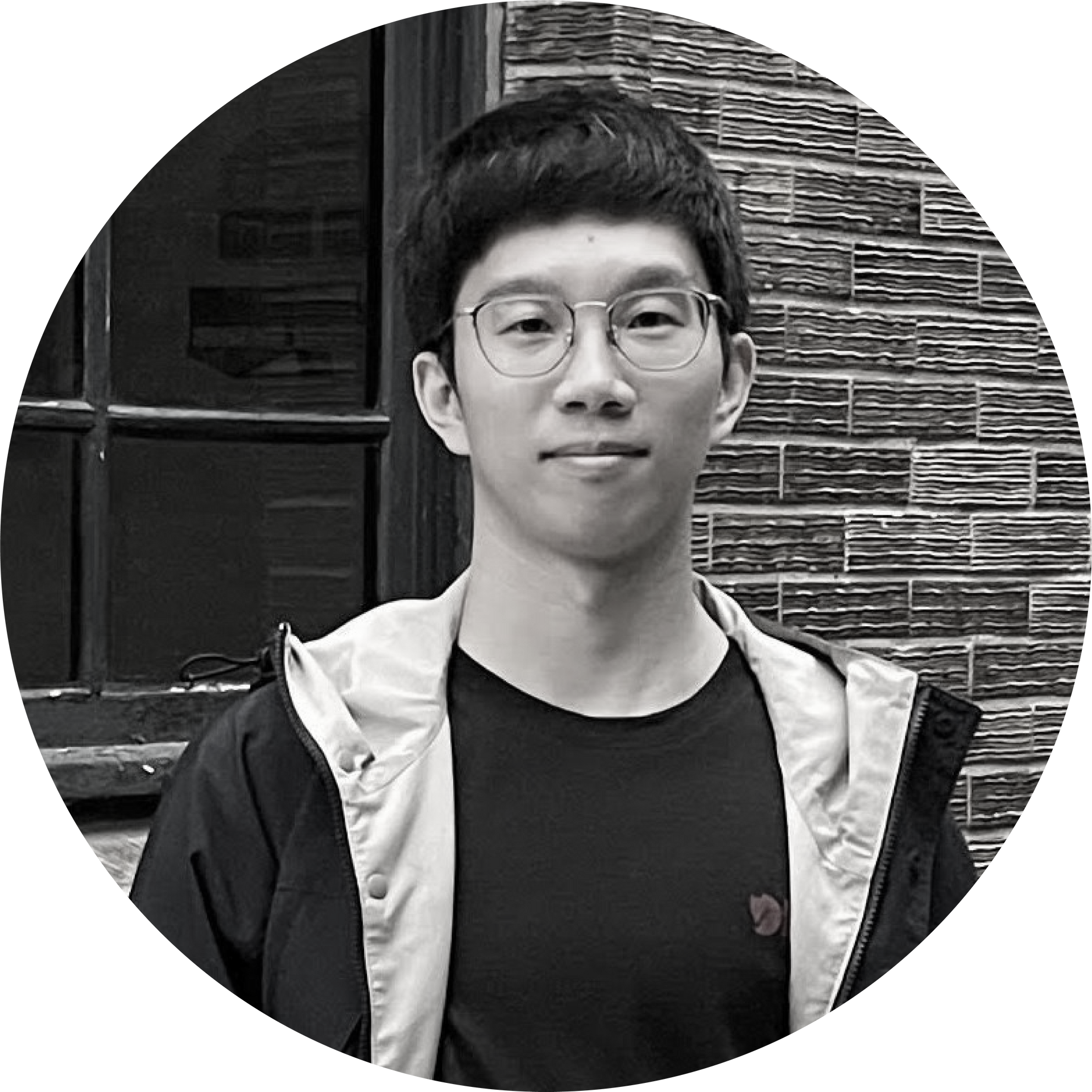 一個戴著眼鏡、穿著夾克的年輕人站在一堵磚牆前。