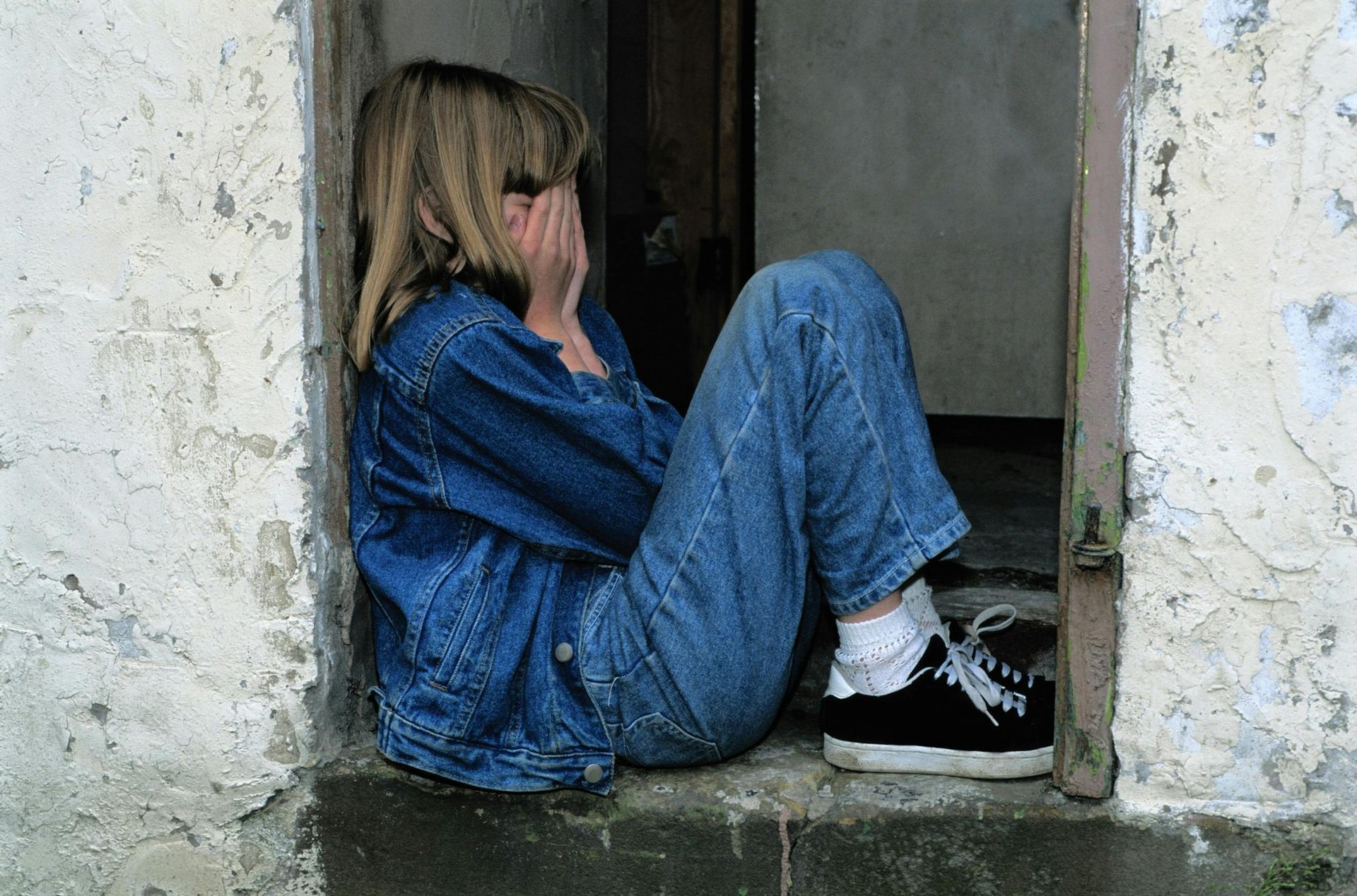 A young girl is sitting in a doorway covering her face with her hands.