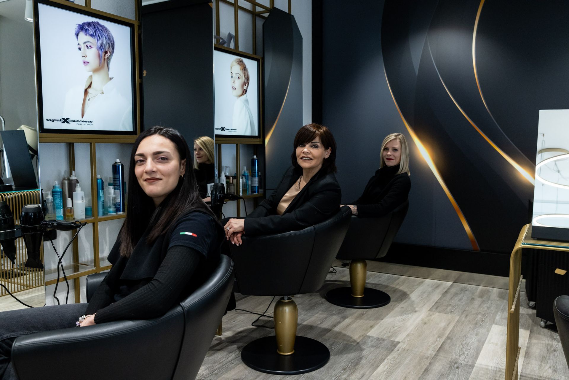 In un elegante salone da parrucchiere nero e oro, tre clienti siedono sulle poltrone.