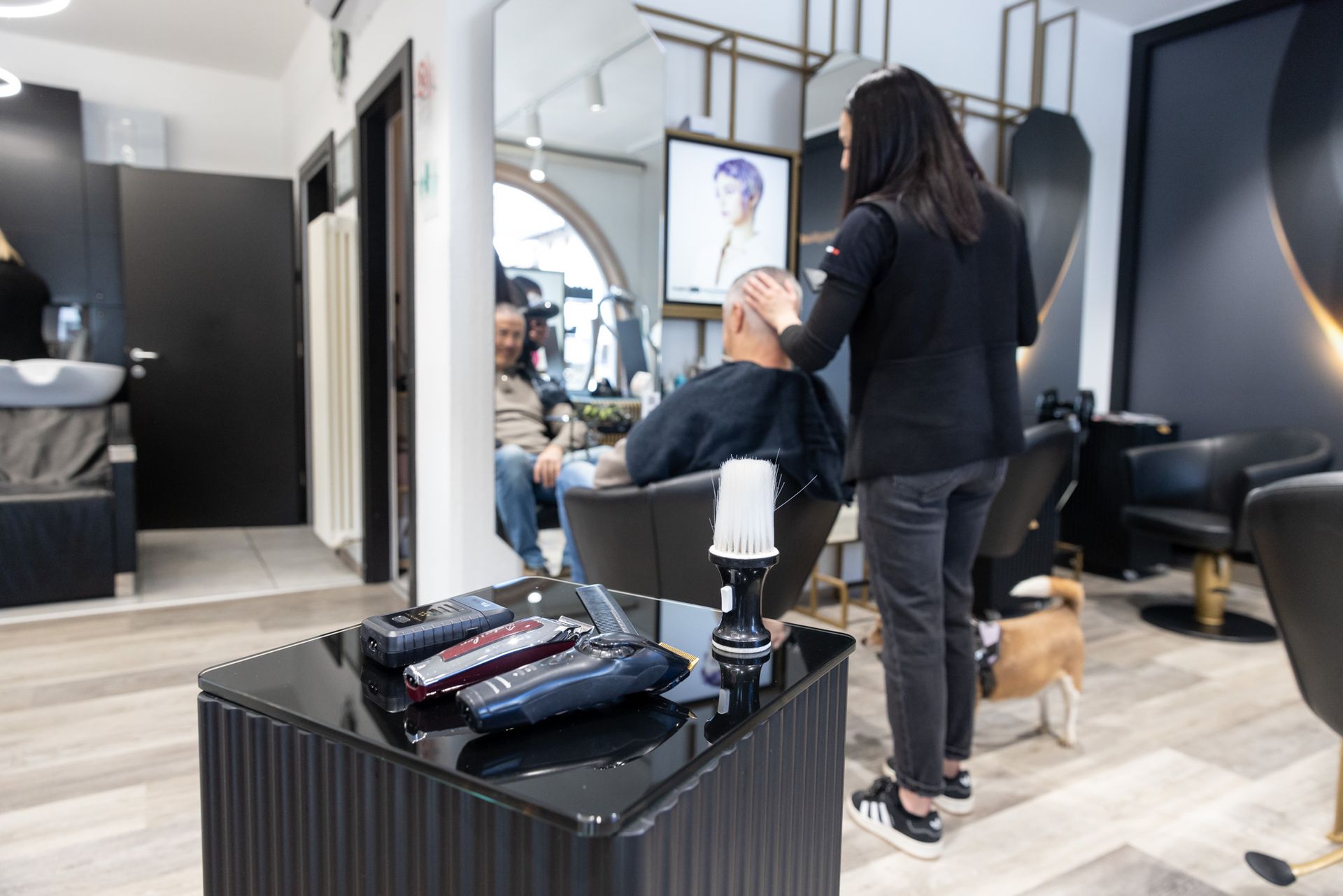 Un parrucchiere asciuga i capelli di una cliente in un salone moderno, con un cane nelle vicinanze.