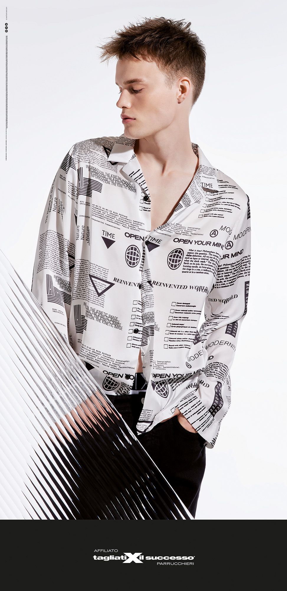 Modello con camicia bianca a fantasia e pantaloni neri, in posa su uno sfondo chiaro.