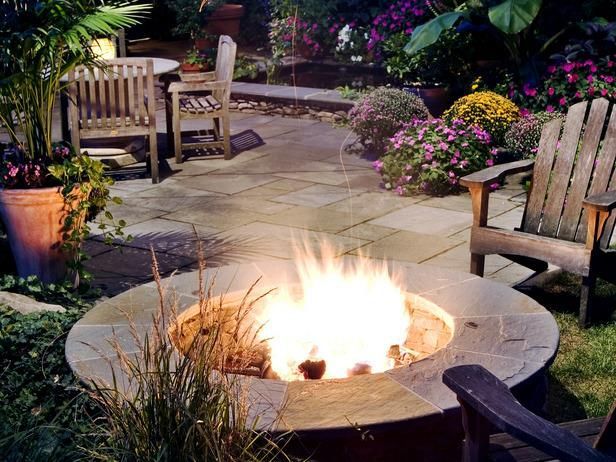 A circular stone fire pit with a bright flame, surrounded by wooden chairs on a patio with plants at dusk.