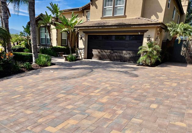 A multi-story house with a tan stucco exterior, dark garage door, and a wide driveway paved with multicolored stone bricks.