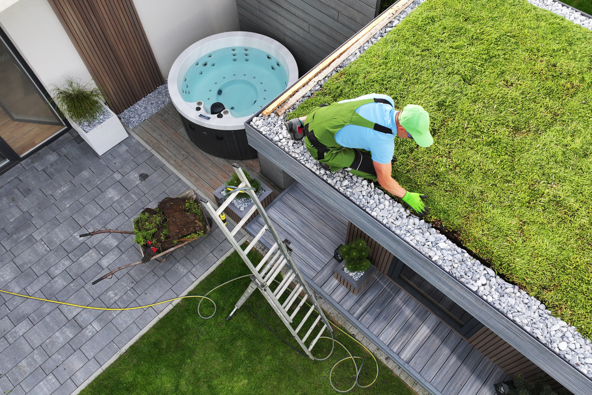 Worker in green clothing trimming a rooftop lawn beside a ladder and hot tub